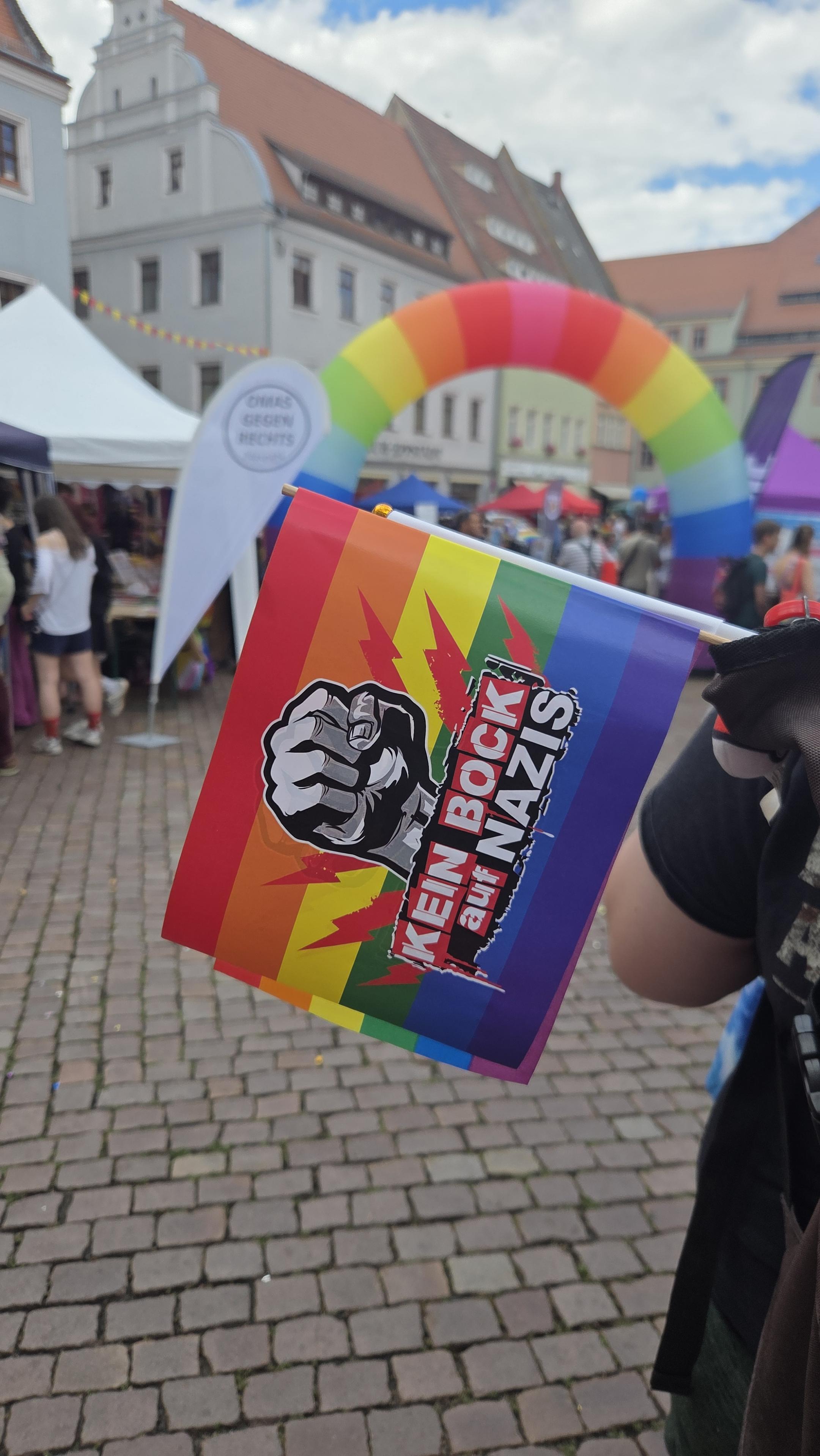 Vor unscharfem Hintergrund eine Papierflagge in Regenbogenfarben mit einer Faust und Aufschrift "KEIN BOCK auf NAZIS"