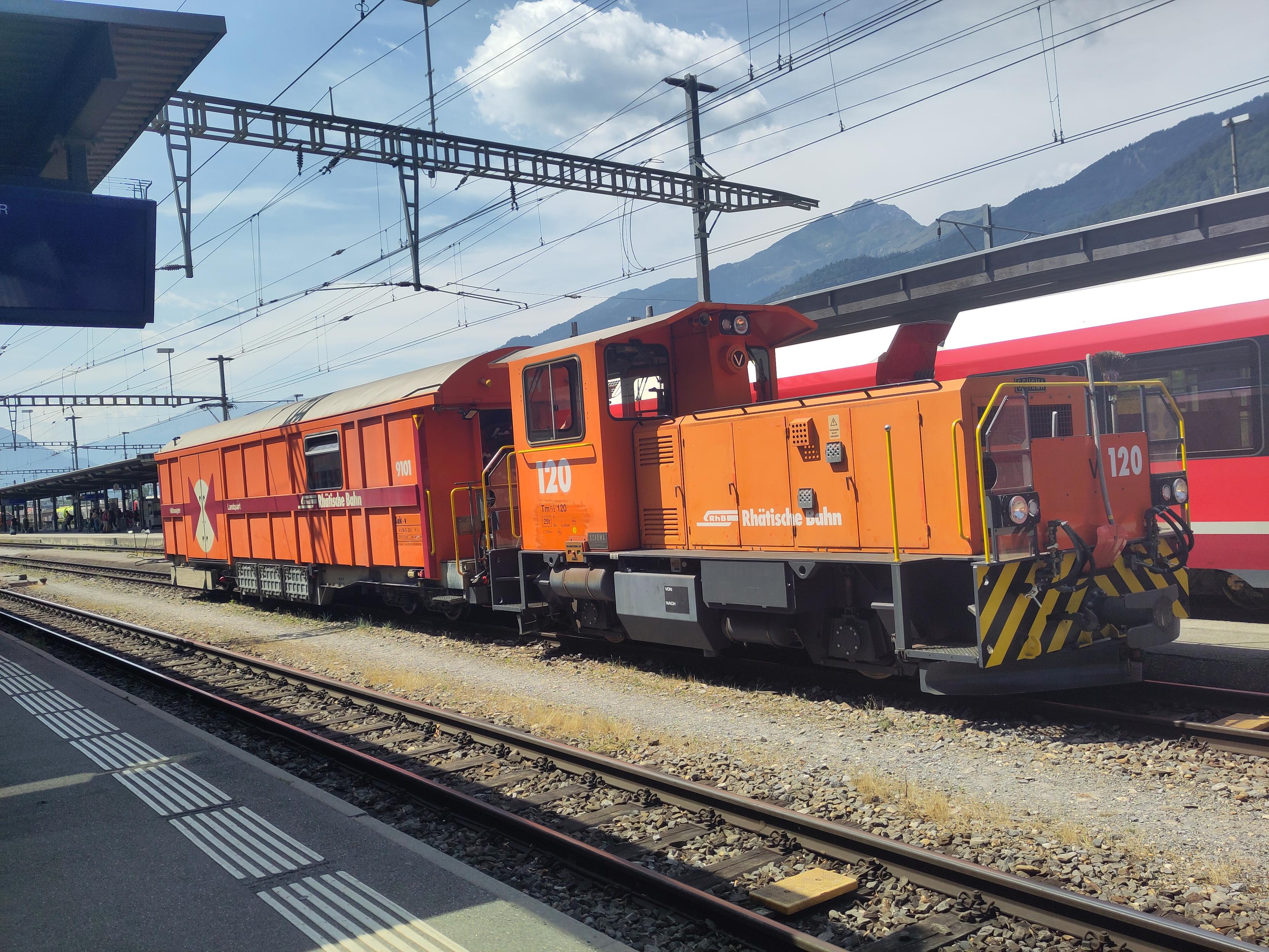 Foto. Eine Rangierlok mit einem orangefarbenen Wagen im Bahnhof.