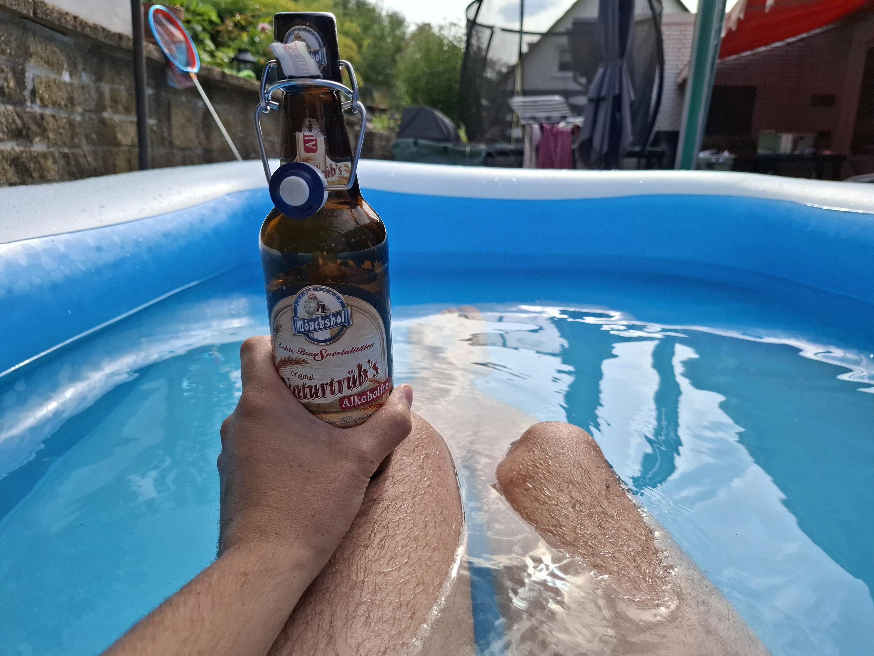 Zwei wirklich markant schöne Beine in einem großen Planschbecken, darauf eine gepflegte Hand am Arm die eine Flasche Alkoholfreies Bier in der Hand hält, die Sonne scheint die Welt ist schön und der Pool (haha Planschbecken) blau
