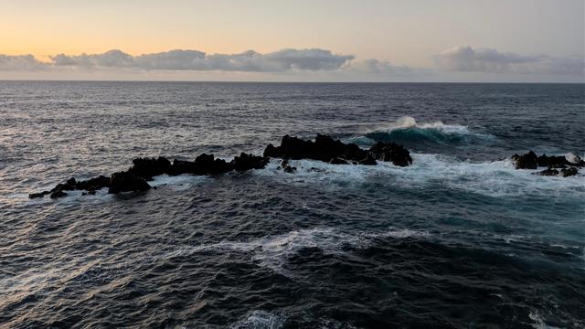 Kleine Felsgruppe im Meer im Restlicht einer untergehenden Sonne, die noch einen Hauch roa an den Himmel zaubert. Ein Wolkenband unterbricht die rosa Färbung. Die schwarze Felsgruppe in der Bildmitte wird von Wellen umspült, auf der rechten Seite türmt sich eine hohe Welle auf,deren Gischt im Gegenlicht spritzt.