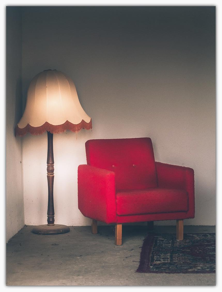 Nahaufnahme einer roten Polstersessel‑Ecke mit hölzernen Füßen, daneben eine hohe Stehlampe mit beigefarbenem Stoffschirm samt gewelltem Rand und Fransen; der Betonboden und die schlichte, helle Wand erzeugen eine minimalistische, leicht vintagehafte Atmosphäre, während ein Ausschnitt eines gemusterten Teppichs rechts unten sichtbar ist.