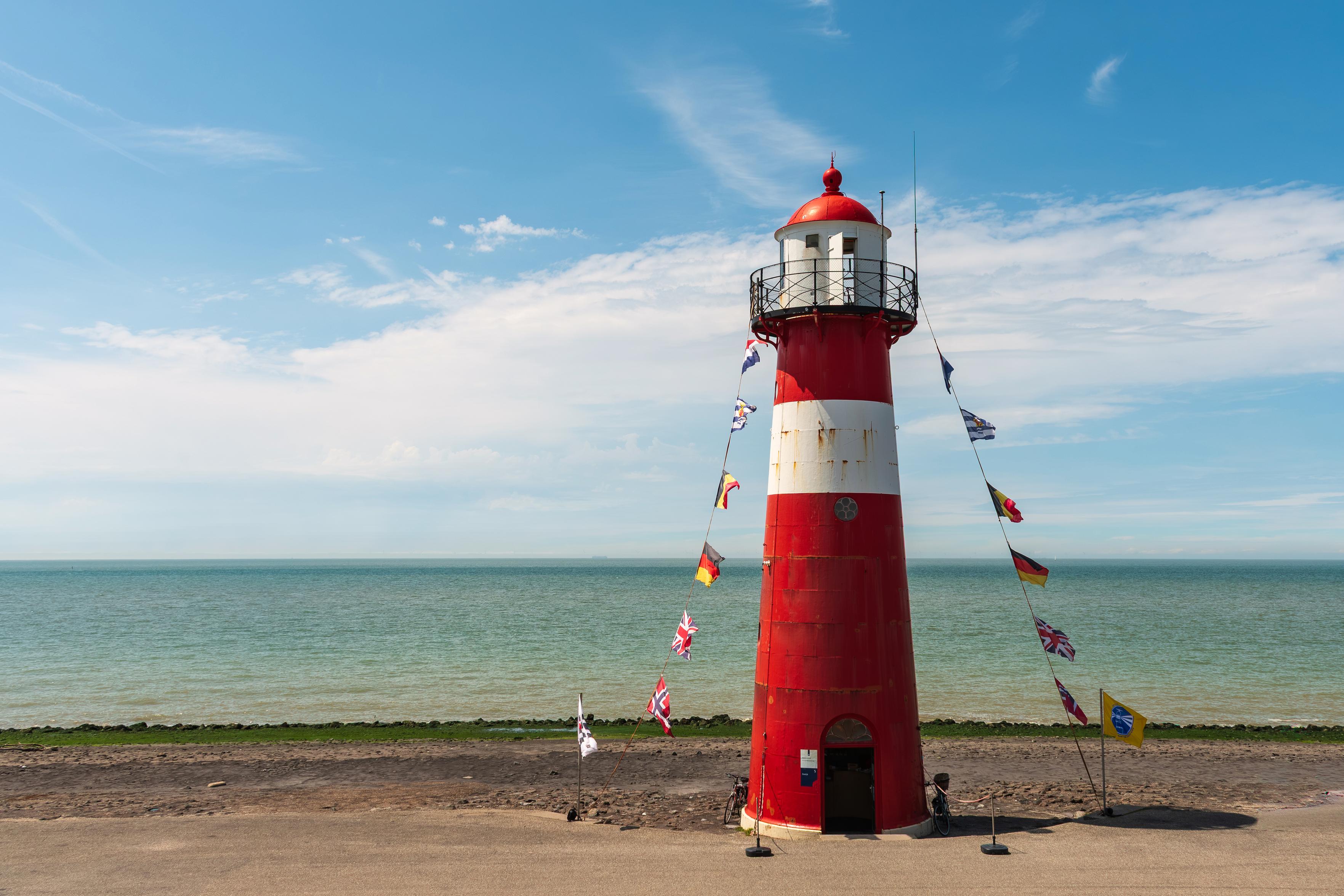 Das Bild zeigt einen roten und weißen Leuchtturm am Meer. Mehrere Flaggen verschiedener Länder sind an Schnüren vom Leuchtturm herabgezogen. Der Himmel ist blau mit einigen weißen Wolken. Das Meer ist ruhig und die Küste ist mit Gras bewachsen.