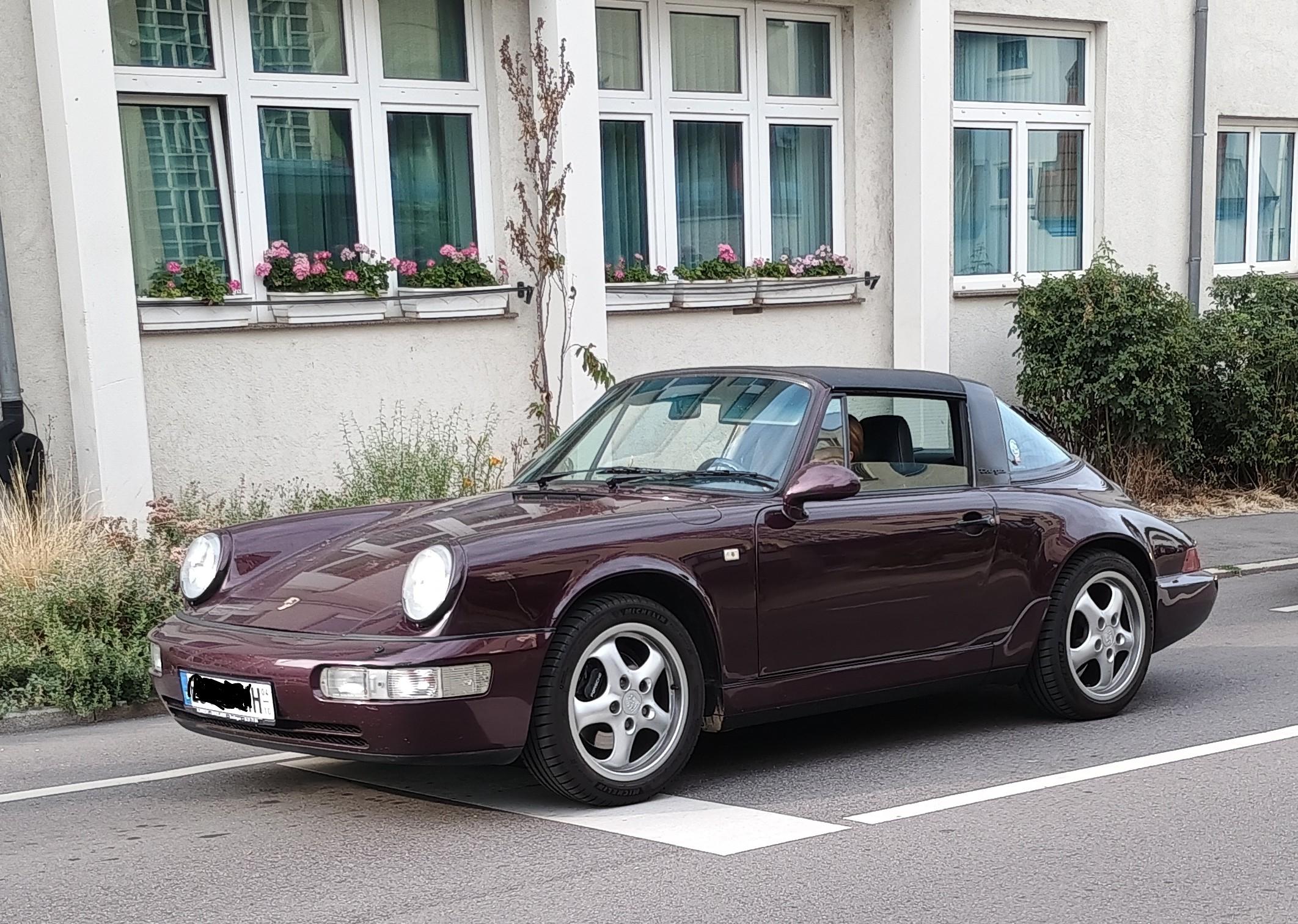 Brauner Porsche mit H im Nummernschild steht vor einem hellen Gebäude. Die Fahrerseite zeigt zur Kamera. Die Vorderräder stehen auf dem Haltebalken, die Frontseite ragt deutlich über den Haltebalken hinaus.