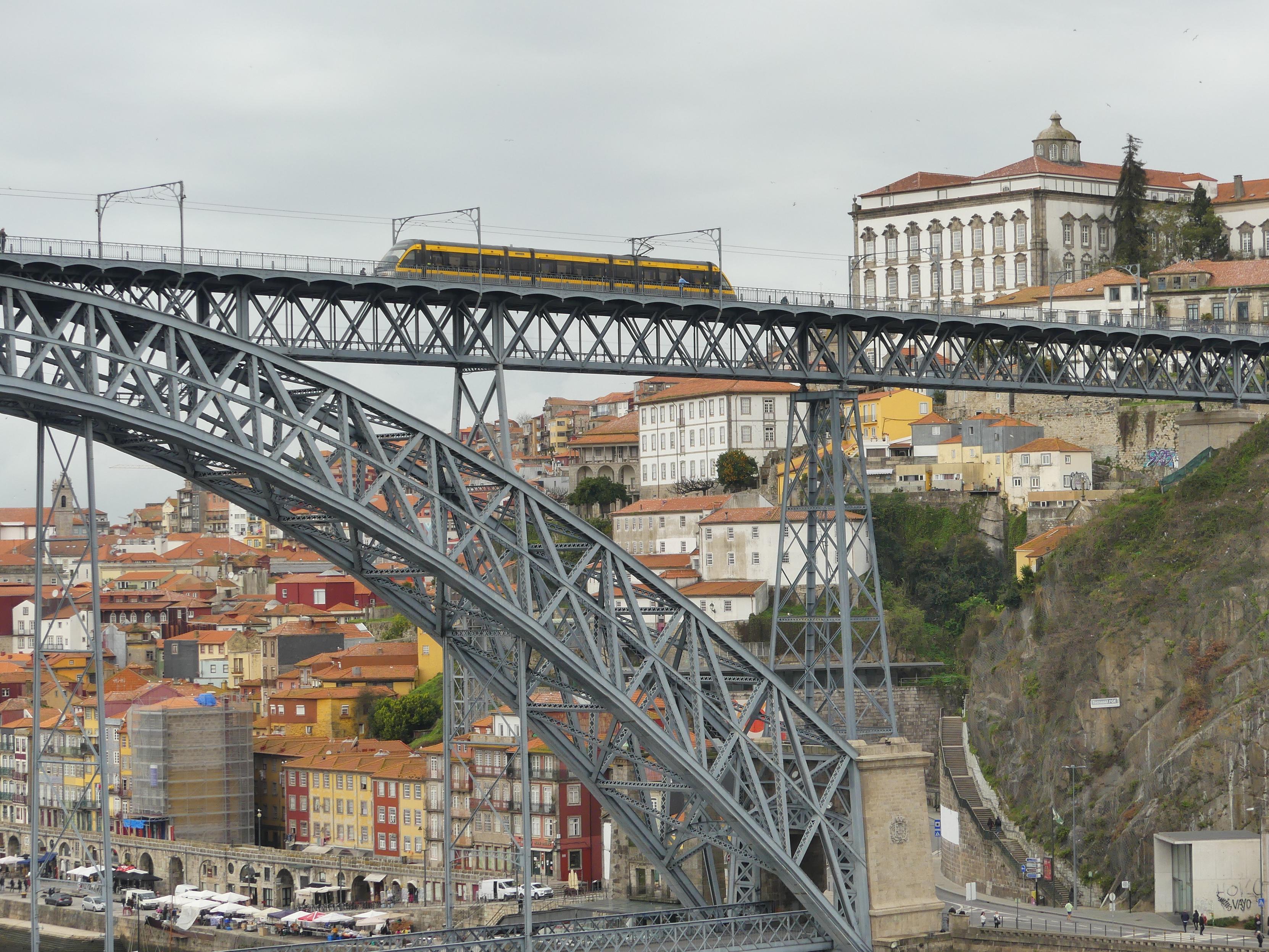 Foto auf die Altstadt von Porto. Im Vordergrund die halbe Brücke über der Fluss Douro. Oben fährt ein Zug der Metro. Dahinter am steilen Hang viele Häuser der Altstadt.
Quelle: eigenes Werk.