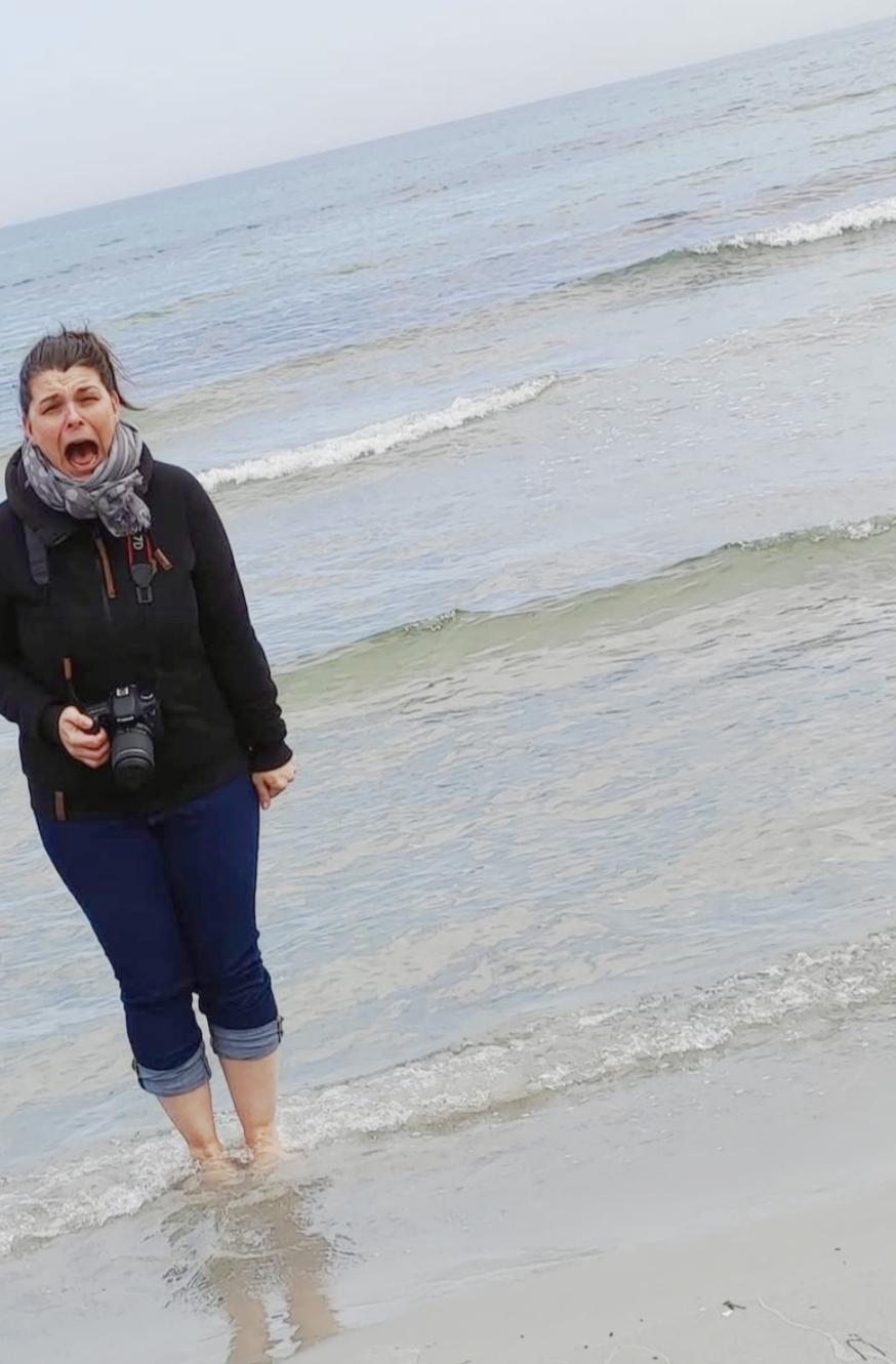Eine Person steht im seichten Wasser an einem Strand. Die Person hält eine schwarze Kamera in der rechten Hand und hat den Mund weit geöffnet. Die Kleidung besteht aus einer dunklen Jacke, einem grauen Schal und dunkelblauen Hosen, die bis unter die Knie hochgekrempelt sind. Kleine Wellen umspülen die nackten Füße der Person. Im Hintergrund erstreckt sich das Meer unter einem grauen, bewölkten Himmel.