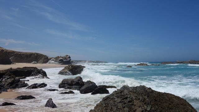 Wellen branden auf einen mit schwarzen Felsen durchsetzten Sandstrand.  Wass türkis, die Wellen brechen in weißer Gischt und sind ca einen halben Meter hoch. Der Sandstrand ist helles ocker, darüber strahlend blauer Himmel ohne Wolken am Mittag.