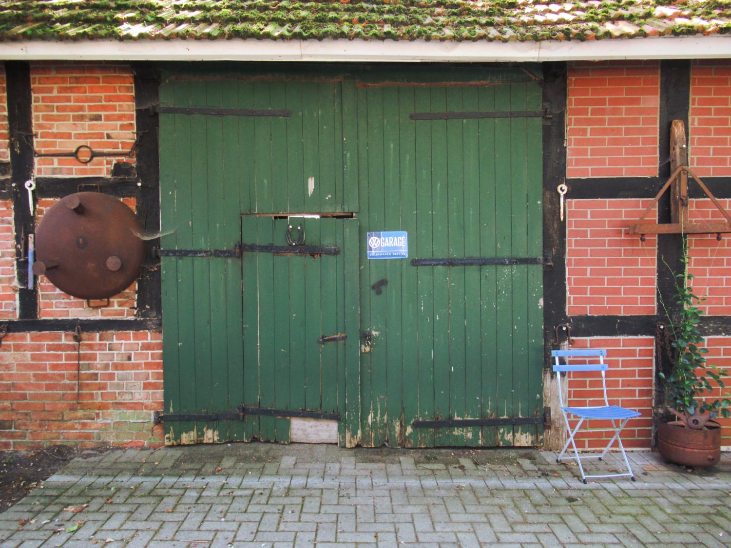 Ein grünes Holztor in einer Fachwerkfassade mit rotem Klinker und schwarzen Balken. Rechts ein hellblauer Klappstuhl und eine Pflanze in einem großen Blumentopf. Links hängt eine Feuerschale an der Wand.
An dem Tor hängst ein Messingschild "Garage – Volkswagen Service"