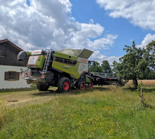 der Mähdrescher steht auf einer Wiese zwischen einem Haus und einem Birnbaum vor dem Haferfeld 