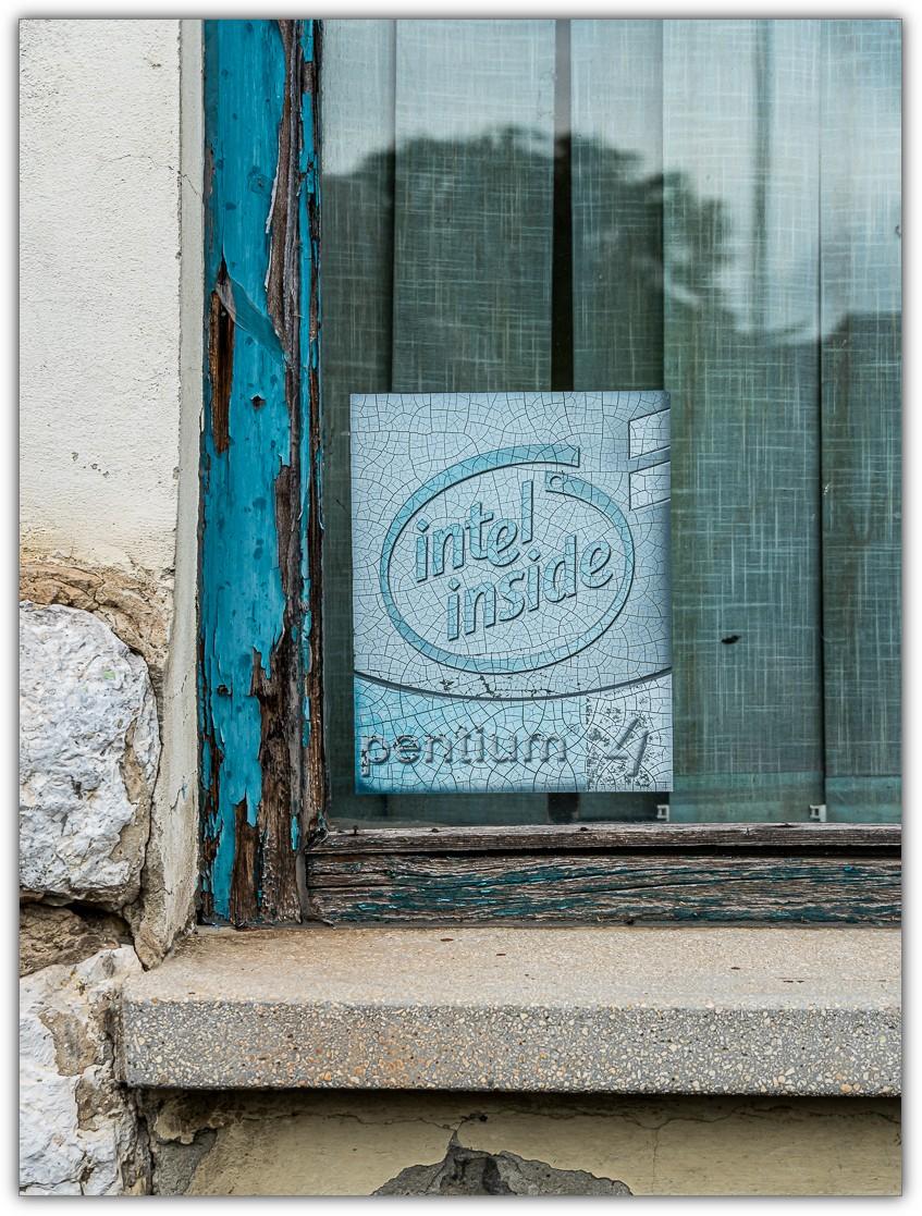 Ein verwittertes Fenster mit abblätterndem türkisfarbenem Holzrahmen und verschmutzter Scheibe. Hinter dem Glas lehnt ein Werbeschild für einen Intel Pentium 4 mit dem Schriftzug „intel inside“. Die Oberfläche des Schildes ist von feinen Rissen überzogen. In der Fensterscheibe spiegeln sich Bäume, während der marode Fensterrahmen und das gealterte Schild zusammen den Eindruck vermitteln, dass hier seit langer Zeit nichts verändert wurde.