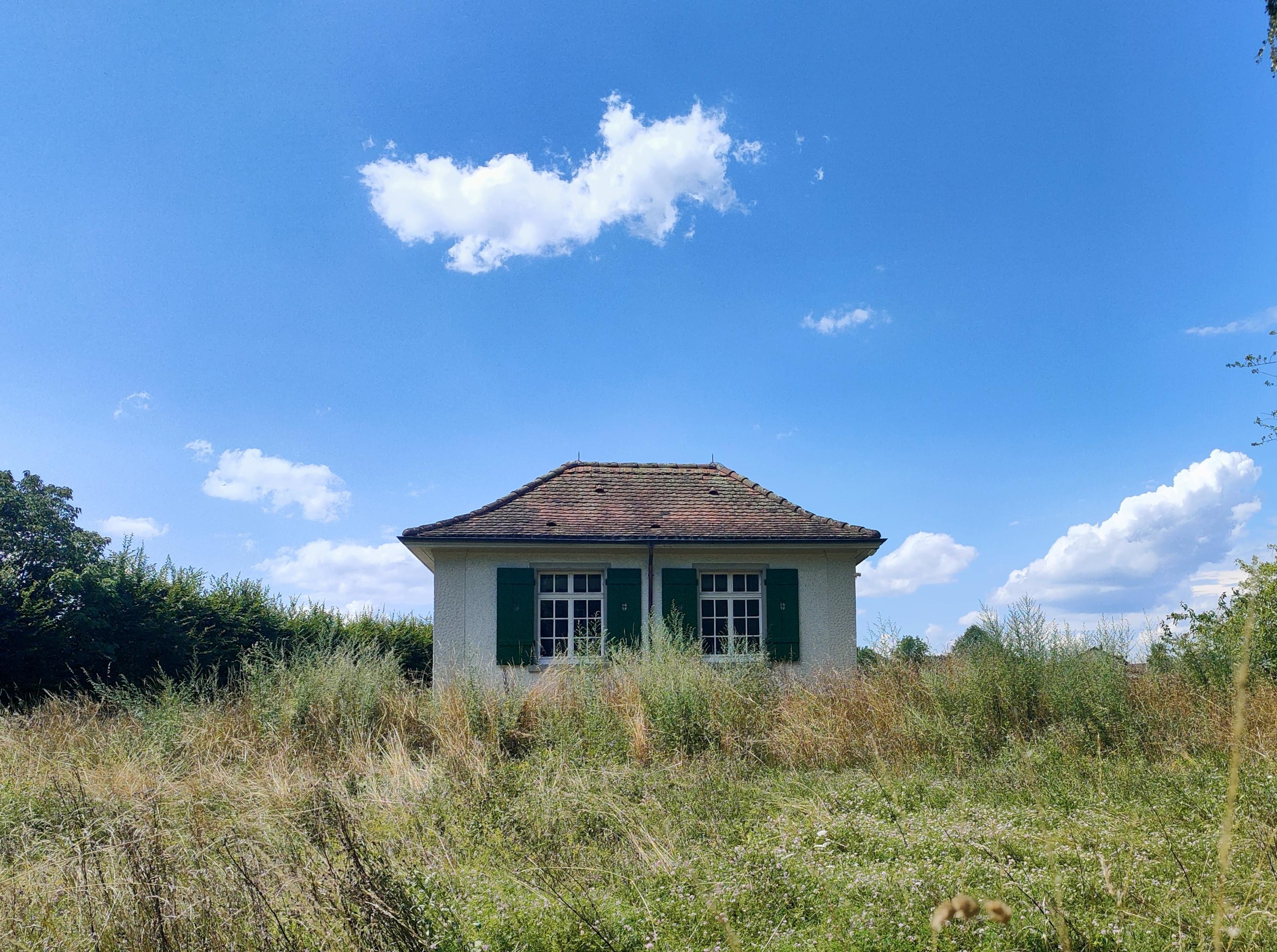 Kleines, einstöckiges Häuschen mit zwei grossen Fenstern an der Front mit grünen Fensterläden und einem kleinen Dach mit braunen Ziegeln.  Man sieht einen blauen Himmel mit Wolken und viel Gras um das Haus herum.