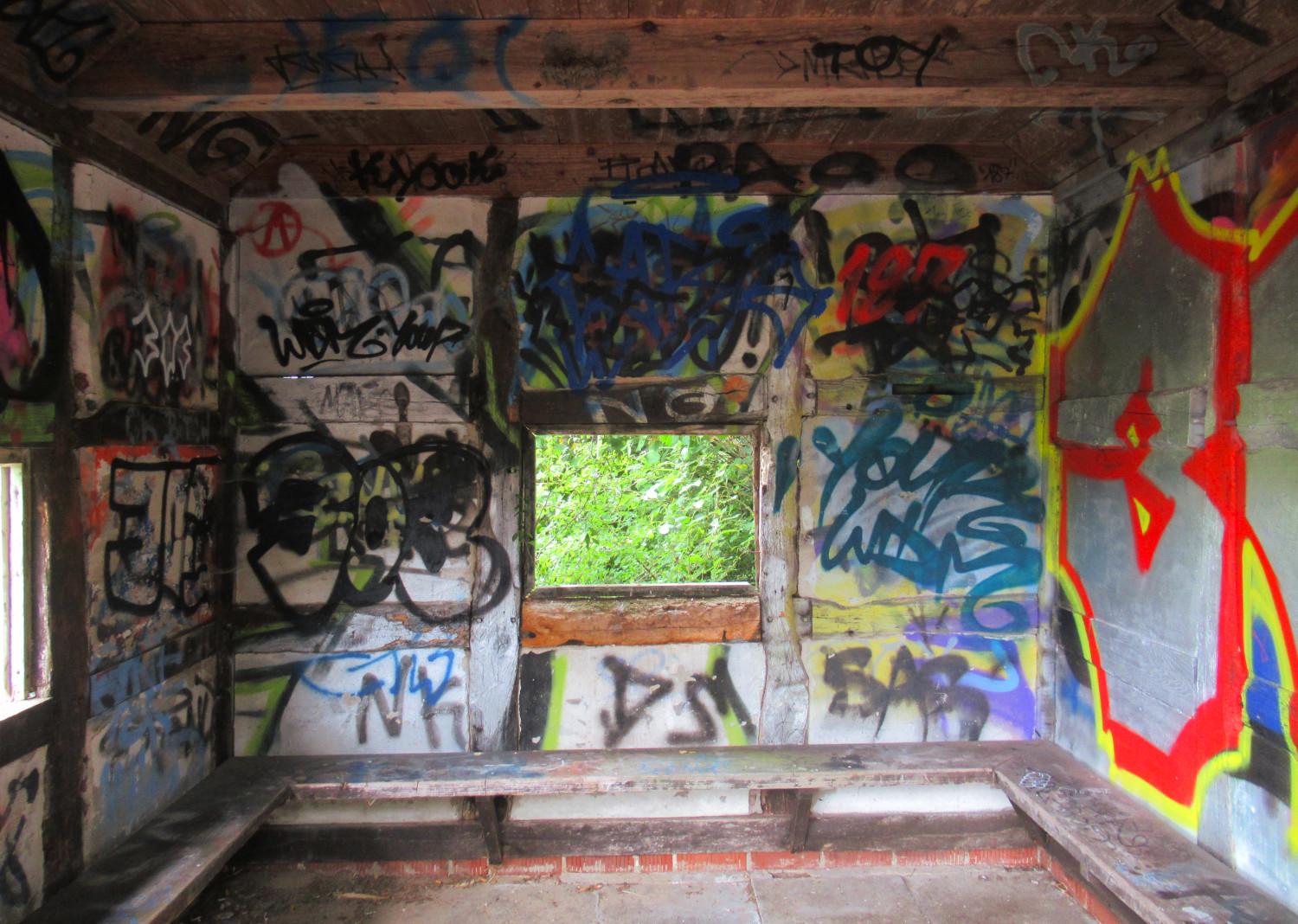 Blick in ein Wartehäuschen aus Fachwerk, ein glasloses Fenster in der Wand, auf die geschaut wird. Die Wände sind mit buntem Graffiti bedeckt.