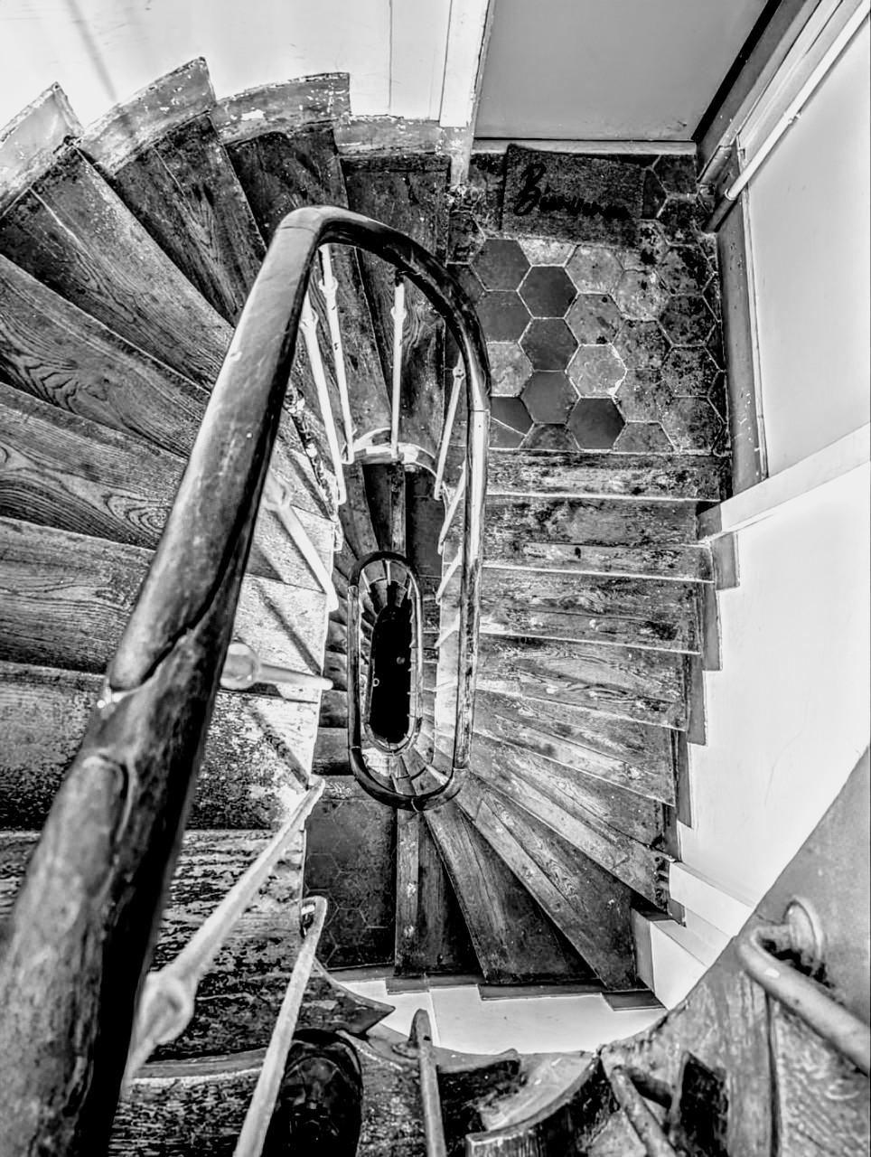 Aufnahme in Schwarzweiß. Ein Treppenhaus, von oben nach unten gesehen.Die Stufen sind aus Holz, alt und abgetreten, ebenso das Geländer. Auf dem Treppenabsatz unter der Fotografin liegt eine Türmatte, auf der "Bienvenue" steht. Das Treppenhaus dreht sich in einer langen Spirale nach unten, das Ende ist nicht zu sehen.