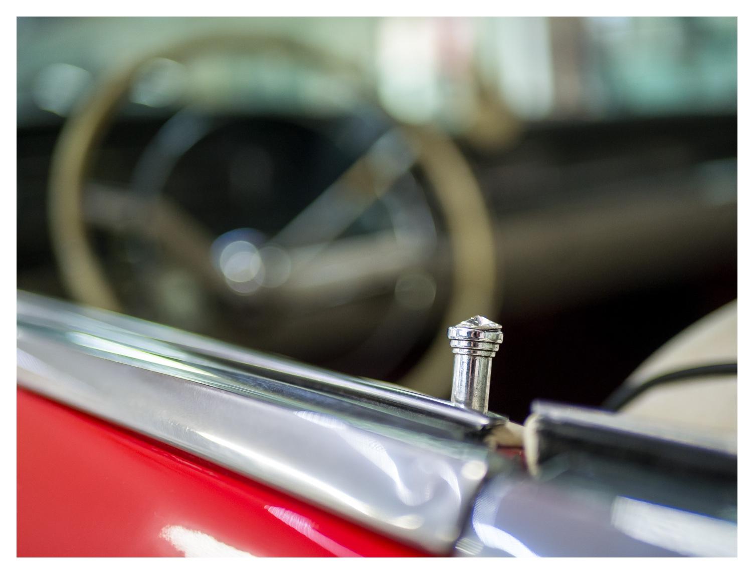 Detailaufnahme der geöffneten Seitenscheibe eines roten Oldtimers. Im Vordergrund steht der verchromte Türverriegelungsknopf scharf im Fokus, während das helle Lenkrad und der Innenraum durch die geringe Schärfentiefe weich verschwimmen. Die glänzenden Chromleisten und der Lack setzen klare Akzente und lenken den Blick auf das kleine Bedienelement.