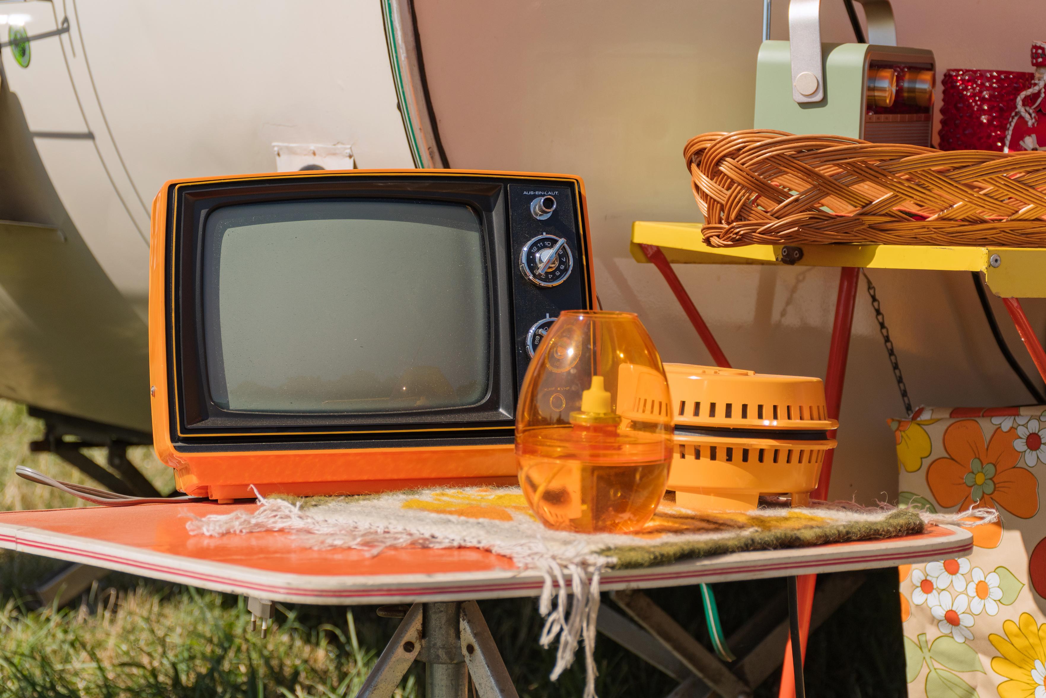 Auf diesem Foto sehen wir eine Ansammlung von Vintage-Gegenständen, die auf einem orangefarbenen Tisch stehen. Im Vordergrund dominiert ein leuchtend orangefarbener Fernseher mit einem dunklen Bildschirm. Daneben steht eine durchsichtige orangefarbene Lampe mit einem gelben Docht und ein weiterer orangefarbener Behälter mit Schlitzen. Diese Gegenstände sind auf einer gemusterten Decke platziert, die über den Tisch gelegt ist. Im Hintergrund ist ein Teil eines Wohnwagens oder Wohnmobils zu erkennen, sowie ein gelbes Regal mit einem Korb und anderen Objekten. Die Szene wirkt wie eine Momentaufnahme von einem Flohmarkt oder einem Campingausflug, der von Nostalgie und dem Charme vergangener Zeiten geprägt ist. Die leuchtenden Farben und die Retro-Objekte erzeugen eine warme und einladende Atmosphäre.