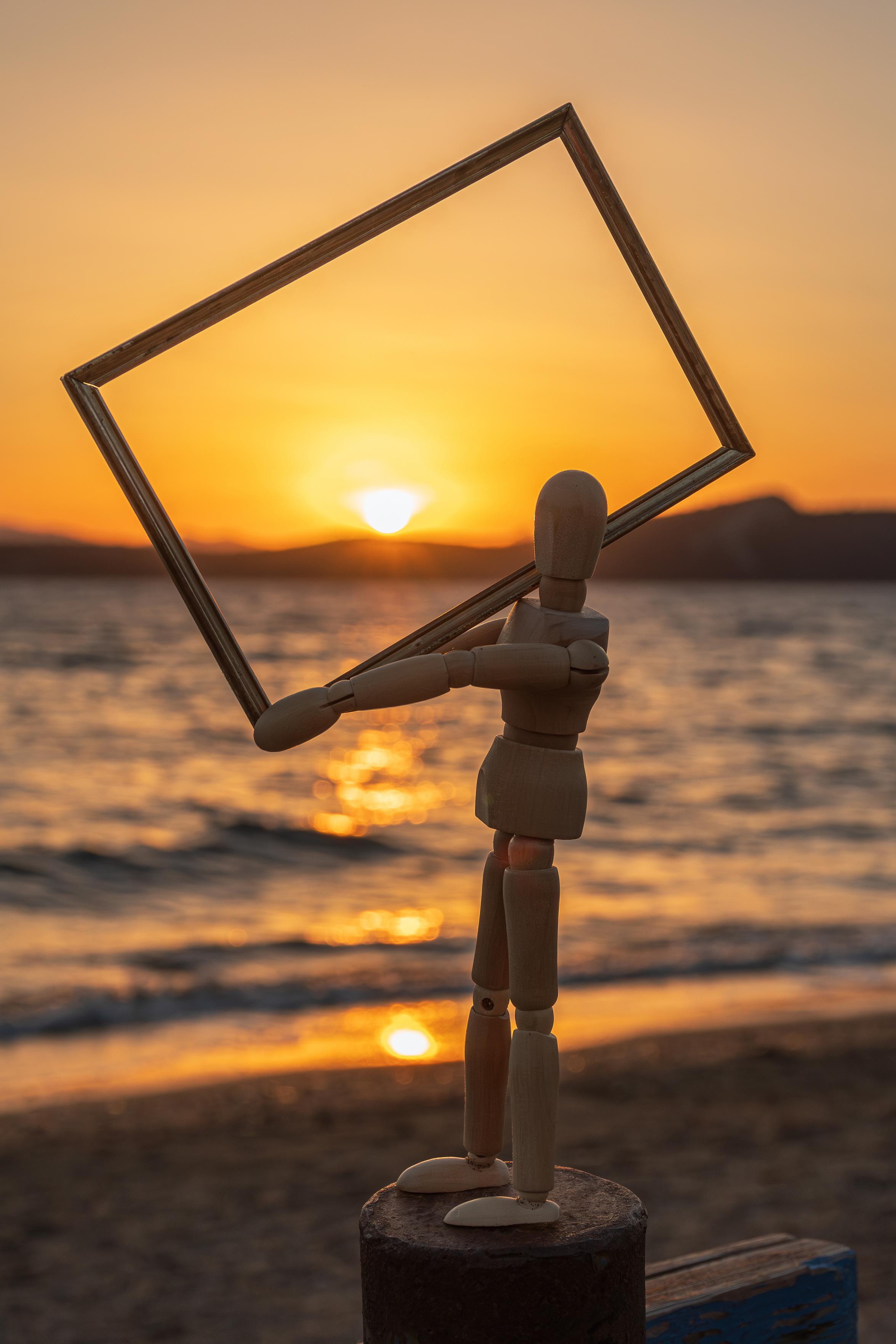 Ein hölzernes Modellpuppe steht am Strand und hält einen leeren Bilderrahmen vor einen wunderschönen Sonnenuntergang über dem Meer. Die Sonne taucht den Himmel in warme Orange- und Gelbtöne, und ihre Reflexion tanzt auf den sanften Wellen.
