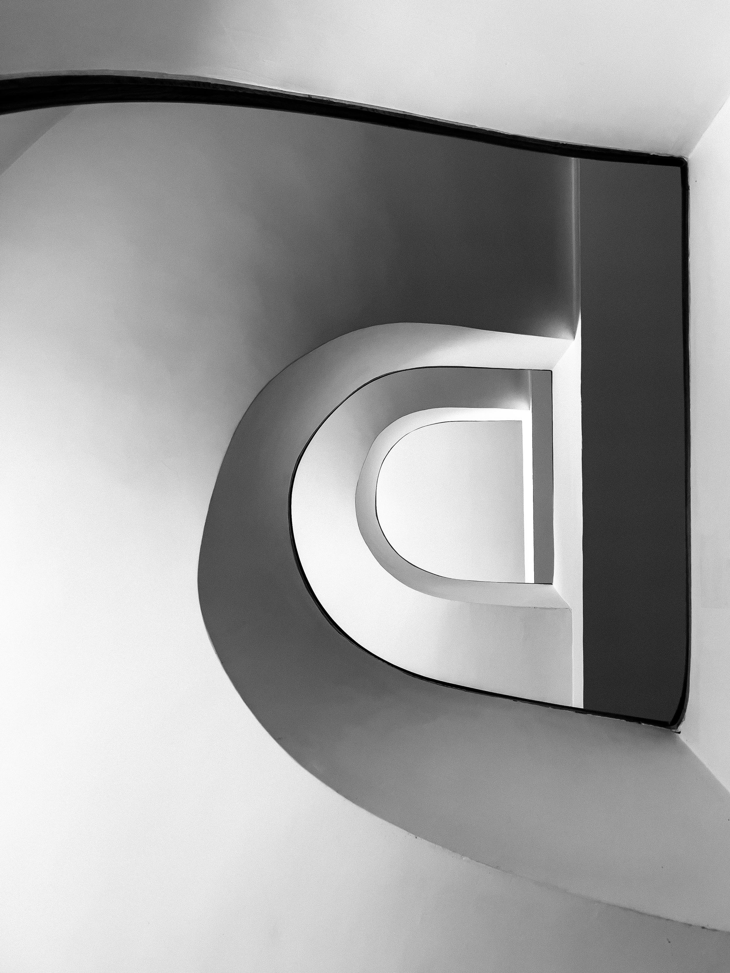 A striking, abstract black and white photograph of a central void in a staircase (the "eye" of a spiral staircase), captured from a low angle. The composition highlights a series of nested, rounded U-shaped forms that recede dramatically into the distance, appearing to be illuminated from within, framing window openings. These central, bright elements are defined by deep, dark, curving shadows cast by the surrounding structure, creating a powerful graphic contour. A dark, rectangular form dominates the top right, framing the composition. The overall aesthetic is highly geometric, minimalist, and sculptural.
