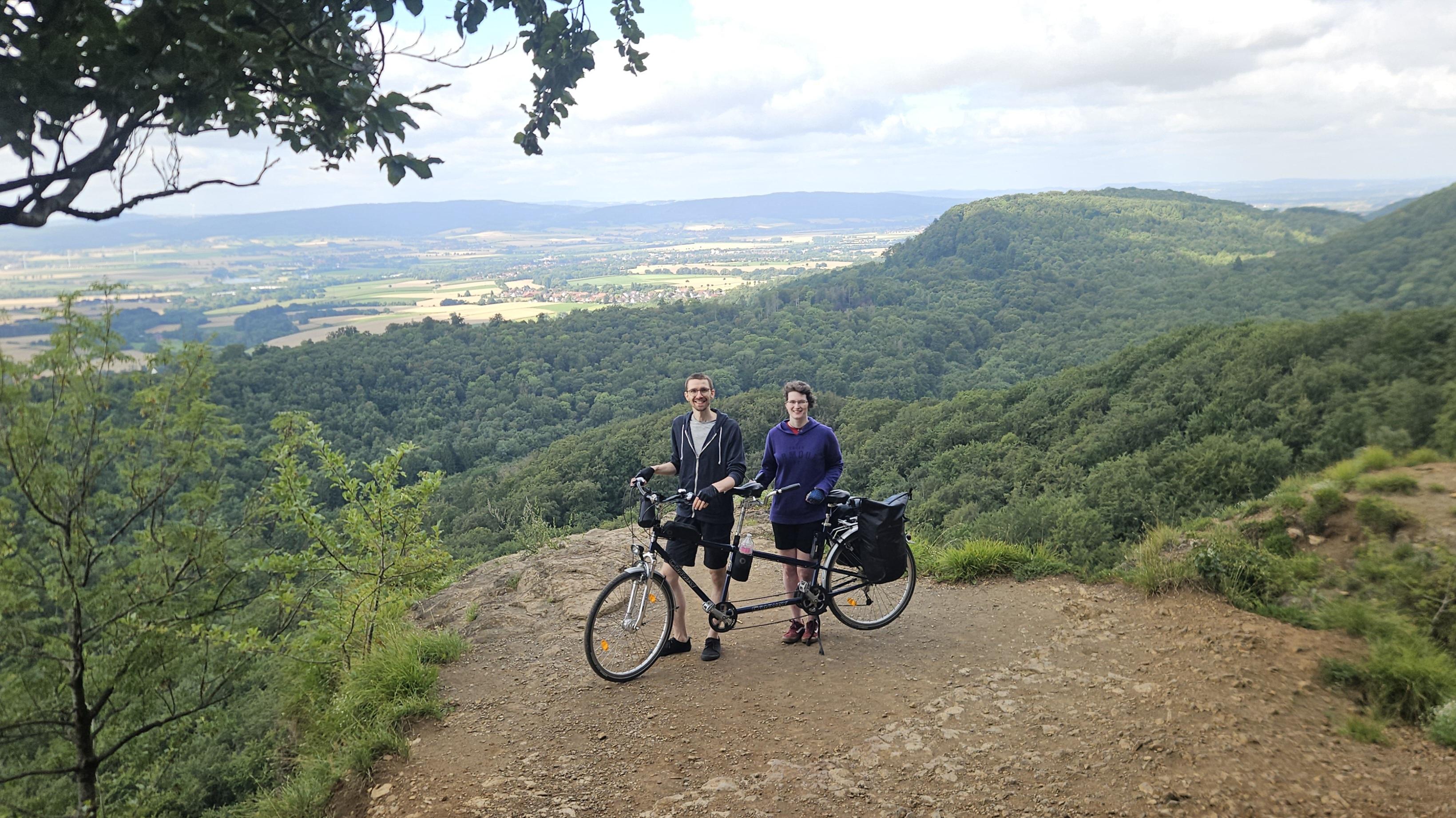 Ein Foto eines Mannes und einer Frau, die beide lächelnd an einem blauen Tandem-Fahrrad auf einem Felsplateau stehen. Im Hintergrund sieht man die Felder, Wiesen und Wälder des Weserberglands.