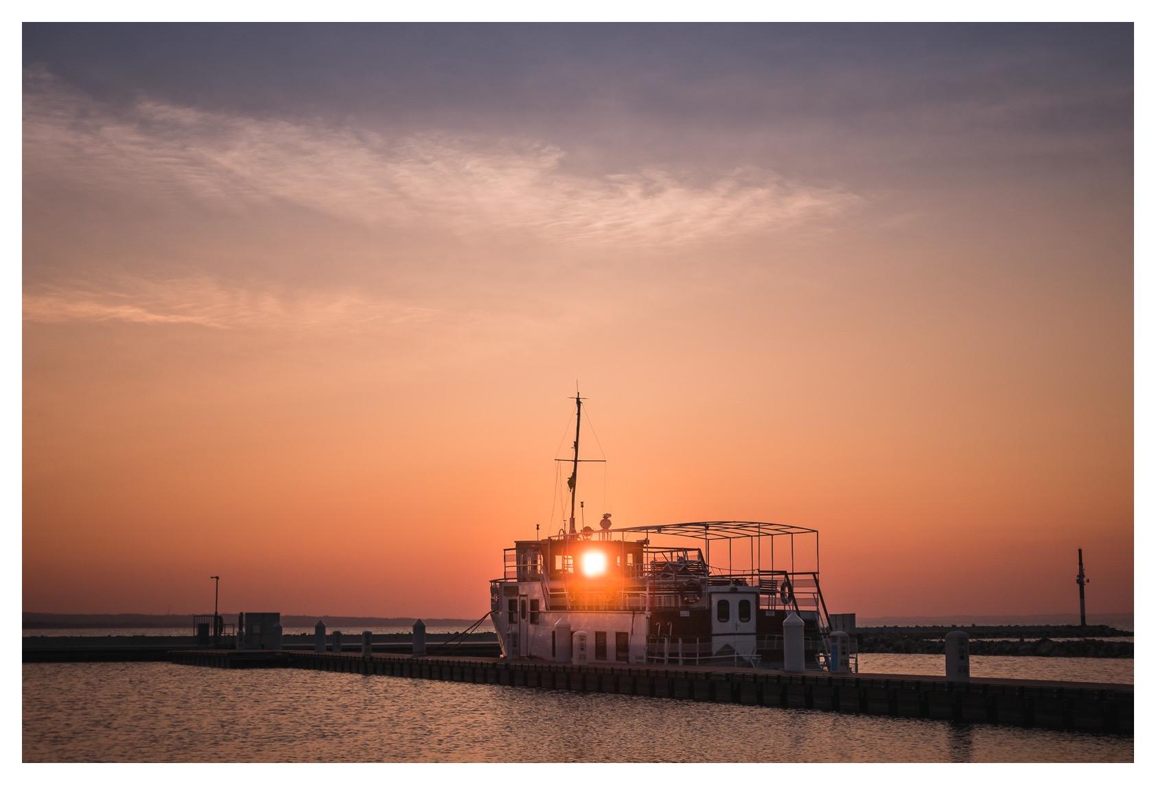 Die untergehende Sonne scheint durch ein Fenster eines am Steg liegenden Ausflugsschiffs und lässt das Schiff teilweise als Silhouette erscheinen. Der Himmel leuchtet in warmen Orange- und Rosatönen und geht nach oben in ein kühles Blau über.