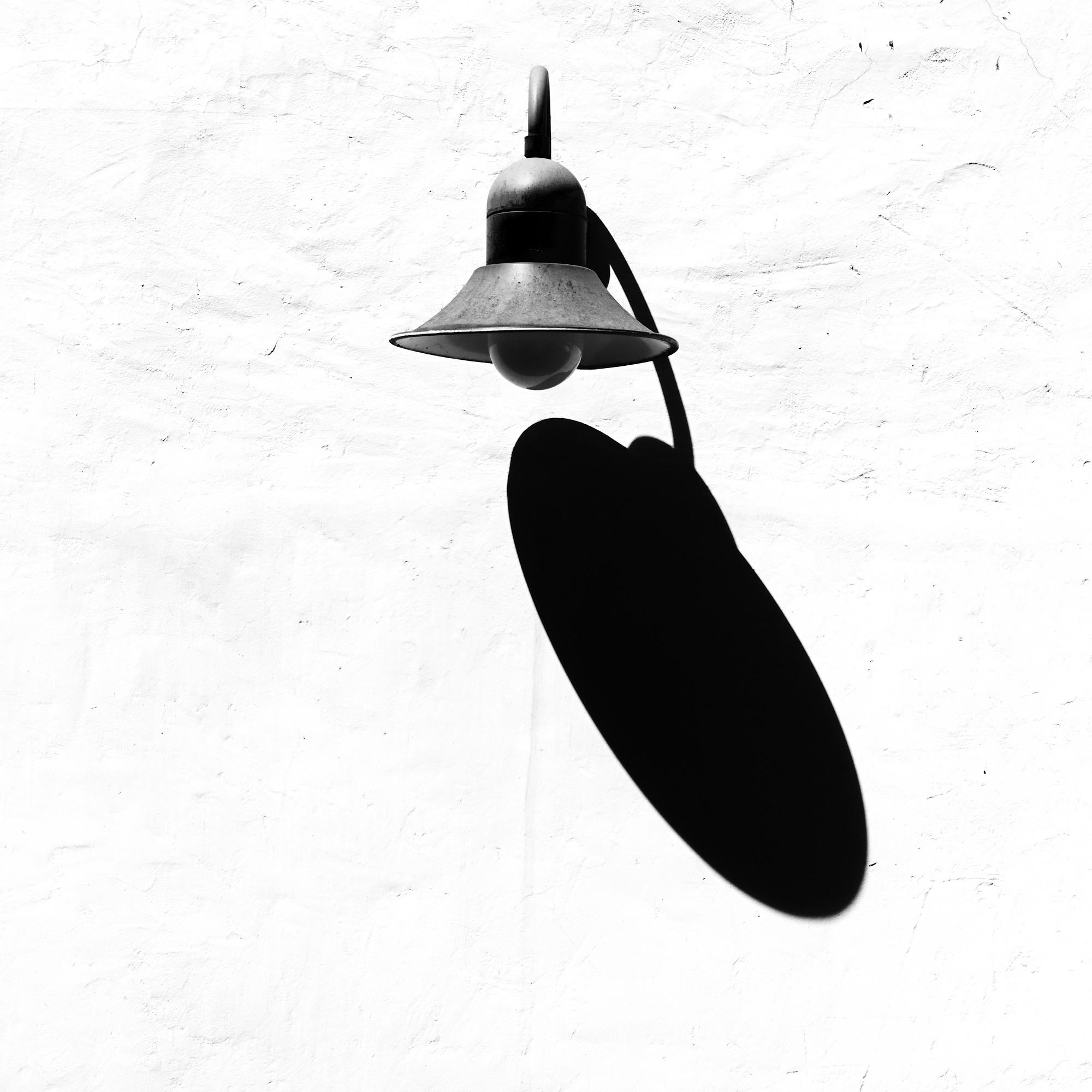 Eine hochkontrastreiche, minimalistische Schwarz-Weiß-Fotografie im quadratischen Format. Eine dunkle, vintage Wandlampe mit metallenem Schirm montiert an einer hellen, leicht strukturierten Putzwand steht im Fokus der oberen Bildhälfte. Das einfallende, harte Sonnenlicht erzeugt einen tiefschwarzen, langgezogenen und perfekt scharf abgegrenzten Schatten, der sich von der Leuchte aus schräg nach rechts unten zieht. Das Wechselspiel zwischen der dreidimensionalen Lampe und ihrem flachen, grafischen Schatten dominiert die schlichte und elegante Komposition.