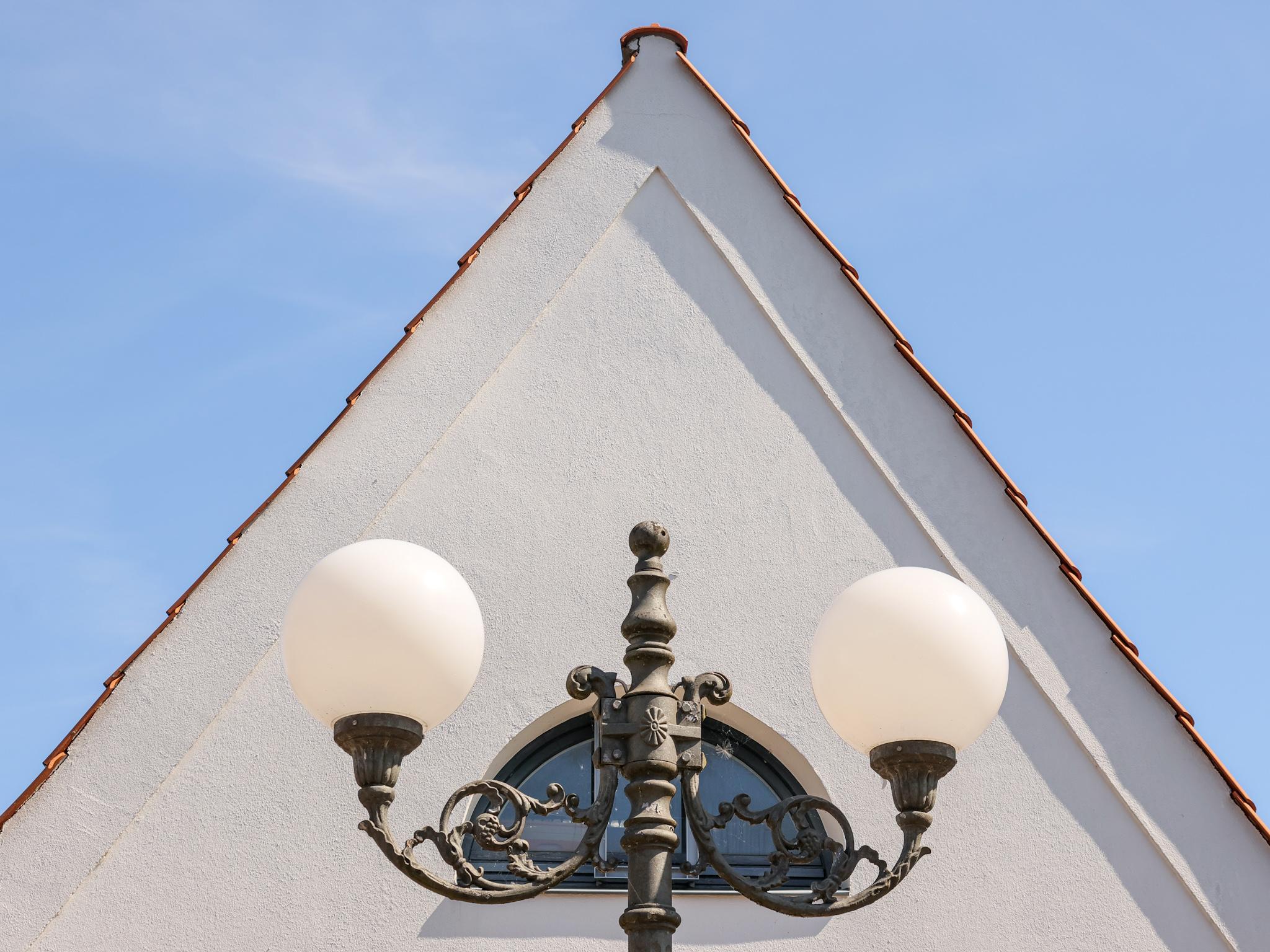 Der weiße Giebel eines Hauses mit halbrundem Fenster. Genau davor eine schmiedeeiserne Straßenlaterne mit zwei Kugelleuchten, welche das Fenster flankieren