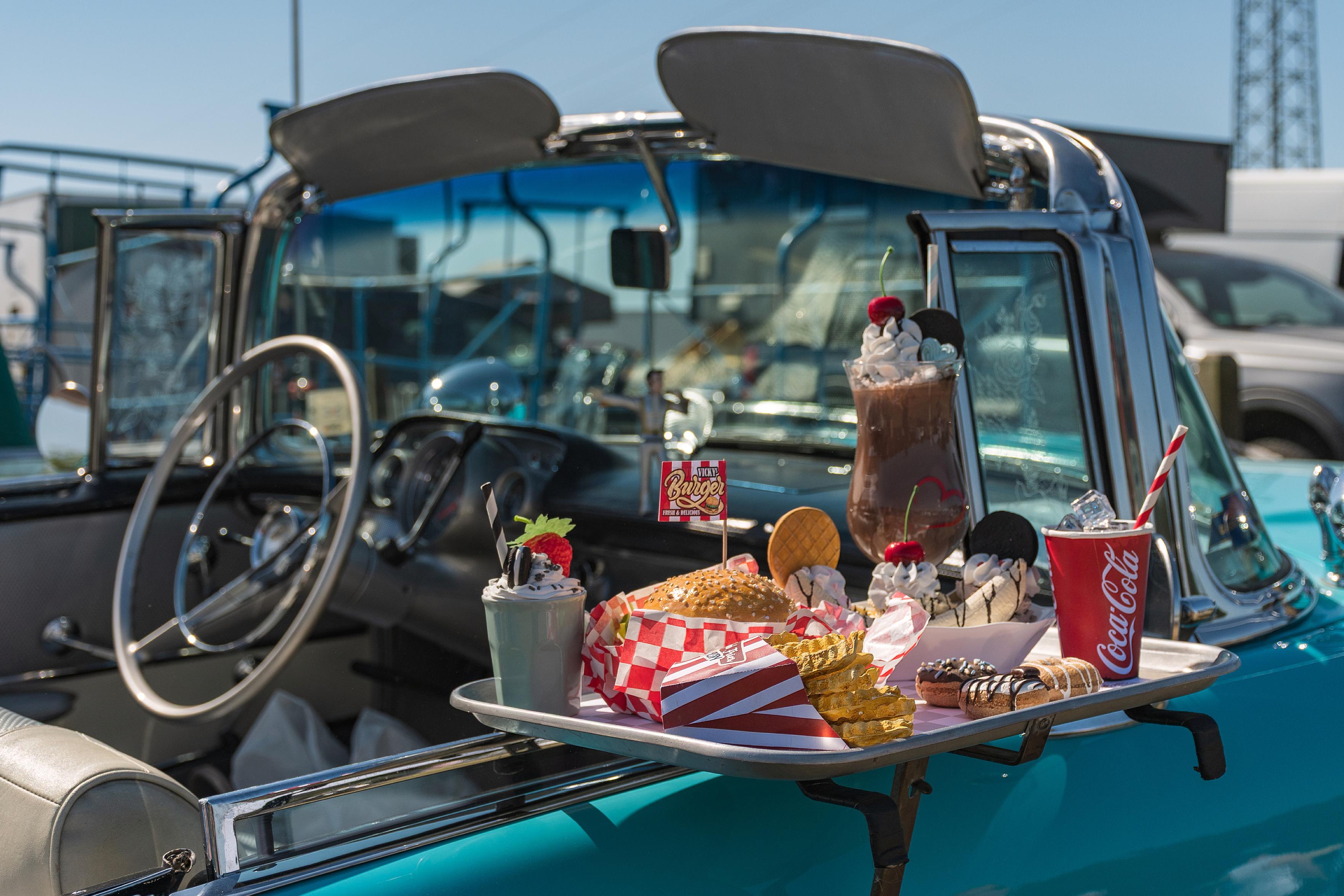 Dieses Foto zeigt eine Szene, die an ein Drive-in-Restaurant erinnert. Ein klassisches, türkisfarbenes Cabriolet steht im Freien, und an der Tür auf der Beifahrerseite ist ein Tablett mit Fast-Food-Speisen platziert. Darauf befinden sich ein Burger, Pommes Frites, ein Schokoladen-Milchshake mit Sahne und Kirschen, ein Becher Coca-Cola mit Strohhalm und einige Donuts. Die Anordnung der Speisen und das Auto deuten auf eine nostalgische Darstellung des amerikanischen Esskultur der 1950er oder 1960er Jahre hin, als Drive-ins und Autos eine zentrale Rolle spielten. Die Szene vermittelt ein Gefühl von Spaß, Genuss und einer Prise Nostalgie.