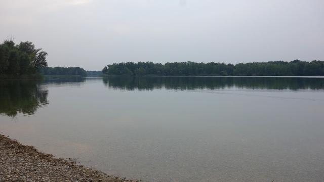 Blick von einem Kiesstrand auf einen See, der Himmel ist stark bewölkt. Keine Schwimmer im Wasser, im See spiegelt sich der Wald auf der anderen Seeuferseite.