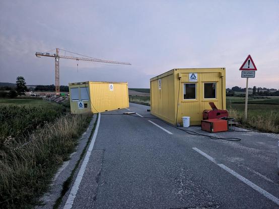 auf einer Straße stehen zwei gelbe Contau.nEiner auf der rechten Straßenseite, vor dem ein umgefallener Stromkasten liegt und ein zweiter gelber Container, der schräg auf der linken Straßenseite und neben der Straße steht 