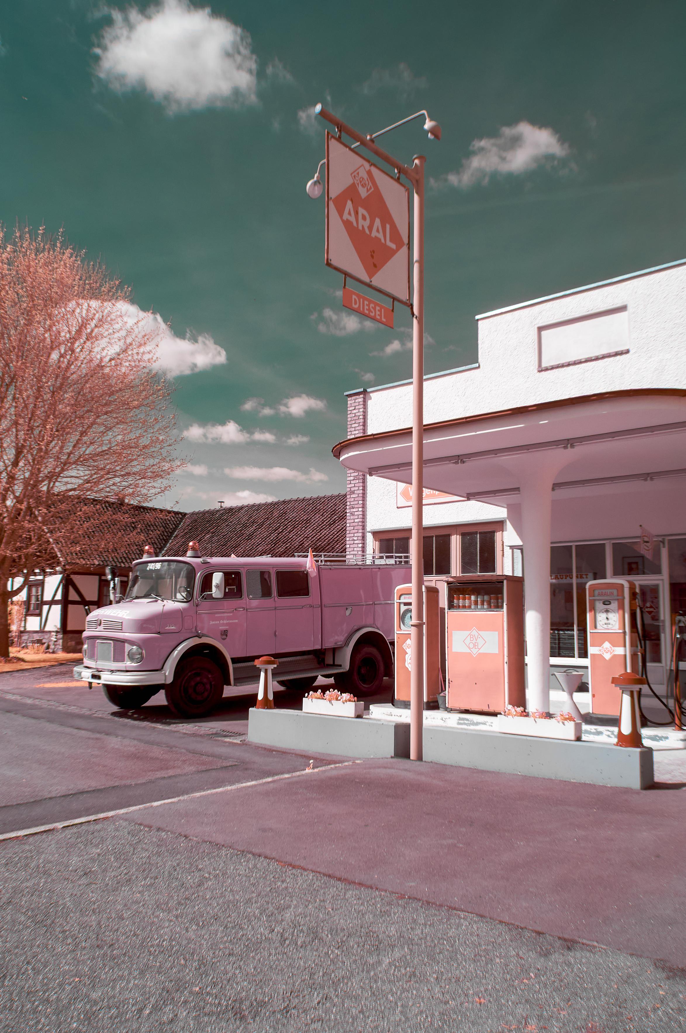 Das Foto zeigt eine Infrarotaufnahme einer nachgebauten Retrotankstelle und ein rosafarbenes Feuerwehrauto, das an der Tankstelle geparkt ist. Die Tankstelle hat eine retro-Architektur mit einem weißen Gebäude und korallenfarbenen Zapfsäulen. Über der Tankstelle hängt ein Schild mit der Aufschrift "ARAL DIESEL". Im Hintergrund ist ein traditionelles Fachwerkhaus zu sehen, und ein Baum mit rosa Blüten steht links im Bild. Der Himmel ist in einem ungewöhnlichen Türkiston gehalten, und die Wolken sind weiß. Die gesamte Szene hat einen leicht surrealen oder verträumten Charakter.