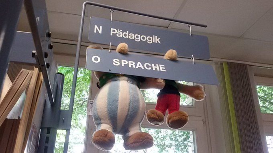Plüschfiguren von Asterix und Obelix, deren Nasen in Hinweisschildern hängen, die offensichtlich in einer Bibliothek auf die Bereiche Pädagogik und Sprache hinweisen.