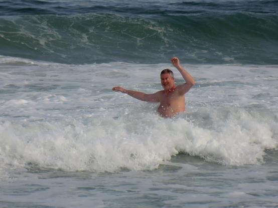 Mann wird gerade von der Brandung einer Welle getroffen und hat die Arme oben. Dahinter türmt sich die Wand der nächste Wasserwand in türkiser Farbe, die auf ihn zurollt.