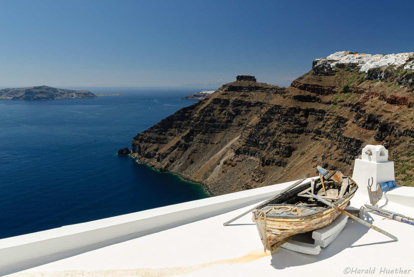 Blick über eine weiße Terrasse auf der links ein altes Ruderboot steht in Santorini. Im Hintergrund die Kippe und das Meer.