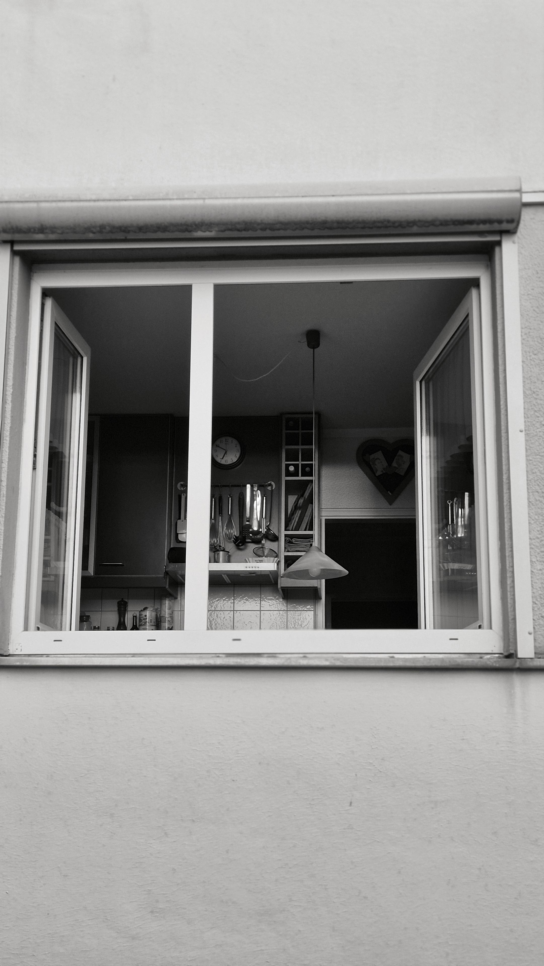 Das rechteckige Hochkantfoto in schwarzweiß zeigt ein geöffnetes Fenster in einem Erdgeschoss von unten, außen aus der Straßenperspektive aufgenommen. Der Fensterrahmen ist weiß, die beiden Flügel stehen nach innen offen. Durch das Fenster ist eine Küche zu erkennen: dunkle Schränke links, eine Uhr an der Wand, hängende Küchenutensilien, ein Regal mit Utensilien und eine Pendelleuchte, die vom der Decken herabhängt. Rechts im Hintergrund ist eine Herzform an der Wand sichtbar. Die Außenwand des Gebäudes hat eine fein strukturierte Oberfläche. Über dem Fenster befindet sich ein Rollladenkasten. Die Aufnahme ist frontal und symmetrisch.

The rectangular, portrait-oriented black-and-white photograph shows an open window on the ground floor, viewed from below from the street. The window frame is white; both sashes are open inwards. A kitchen can be seen through the window: dark cupboards on the left, a clock on the wall, hanging kitchen utensils, a shelf with utensils and a pendant light h