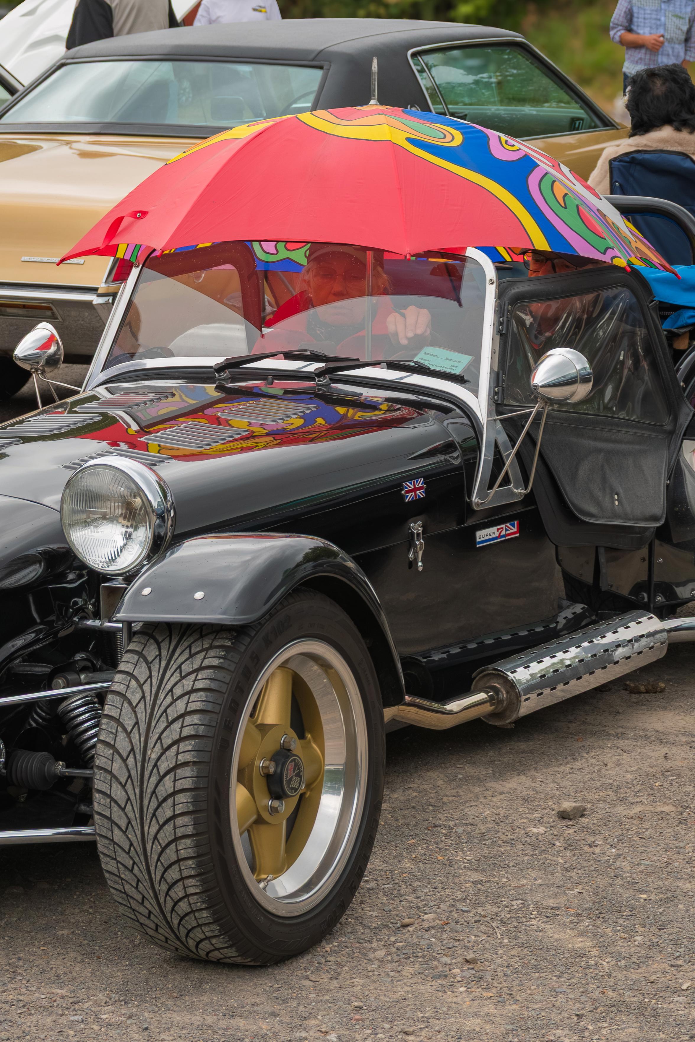 Das Foto zeigt einen historischen schwarzen Sportwagen, der mit einem bunten Regenschirm abgedeckt ist. Der Regenschirm hat ein psychedelisches Muster in Rot, Blau, Gelb und Grün. Er ist über das offene Dach des Autos gespannt und scheint als Sonnenschutz zu dienen. Im Inneren des Autos ist eine Person zu sehen, die hinter dem Lenkrad sitzt und durch den Regenschirm hindurchschaut. Der Wagen hat einen auffälligen Auspuff und goldene Felgen. Im Hintergrund sind andere Autos und Personen zu erkennen, was auf eine Veranstaltung oder ein Treffen hindeutet. Die Szene vermittelt eine spielerische und unkonventionelle Atmosphäre, die durch die ungewöhnliche Verwendung des Regenschirms unterstrichen wird.