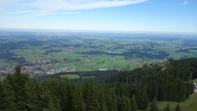Blick von einem Berg in die Ebene und ein kleine Stadt. Die Ebene besteht aus offenen Flächen wie grüne Wiesen, kleinen Wäldchen und die blauen Tupfer sind Seen in der Landschaft. Der Himmel ist blau mit einem blauen Keil von rechts in die weißen Wolken von links.