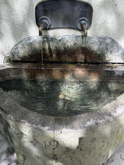 Blick auf einen kleinen Brunnentrog an der Mauer einer Brunnenstube. Er wird von zwei Hähnen gespiesen. Der linke, der sichtbar mehr Wasser speist, ist mit „Seewasser“ angeschrieben. Der rechte, eigentlich nur noch ein Getröpfel, mit „Quellwasser“.