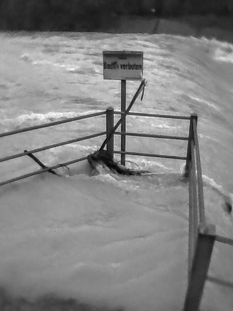 Aufnahme in Schwarzweiß. Ein überspülter Steg in die aufgewühlte Isar mit einem Schild, auf dem steht Baden verboten. Landeshauptstadt München. Der Steg istit einem Geländer aus Metall eingefasst. Am Geländer hängen Algen. Hinter dem Geländer stürzt das Wasser eine Kaskade hinab.