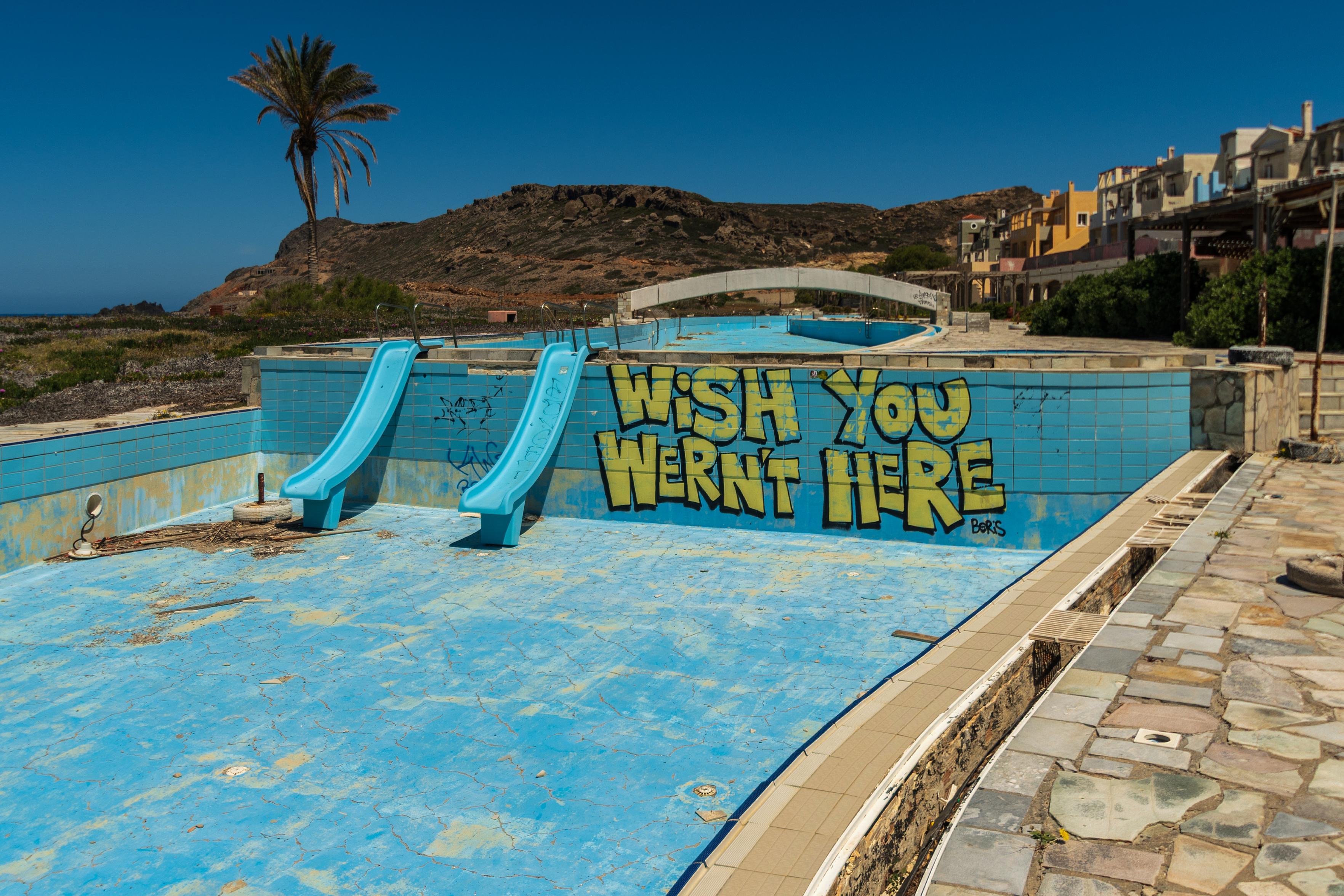 Das Foto zeigt ein verlassenes Schwimmbad, das mit Graffiti übersät ist. Die leeren Becken und die verblassten Farben vermitteln ein Gefühl der Verlassenheit und des Verfalls. Die Graffiti, insbesondere die Botschaft "WISH YOU WERNT HERE", verstärken die melancholische und desillusionierte Stimmung. Die Palmen und die umliegende Landschaft deuten auf ein warmes Klima hin, was den Kontrast zwischen der einst lebendigen Anlage und ihrem jetzigen Zustand noch verstärkt. Es ist ein Bild, das zum Nachdenken über Vergänglichkeit, verpasste Gelegenheiten und die Spuren anregt, die wir hinterlassen.