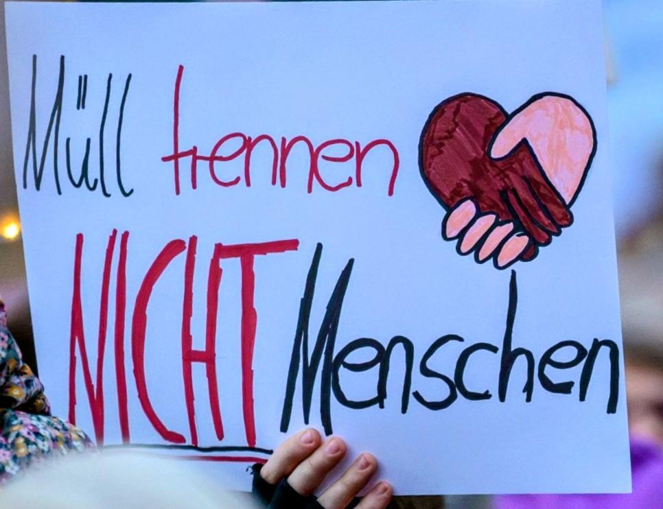 Ein Pappschild auf einer Demo mit der Aufschrift "Müll trennen, NICHT Menschen"
Eine Zeichnung: eine schwarze und eine weiße Hand geben sich die Hand. 
Das Pappschild wird von einer Hand mit fingerlosem Handschuh gehalten.