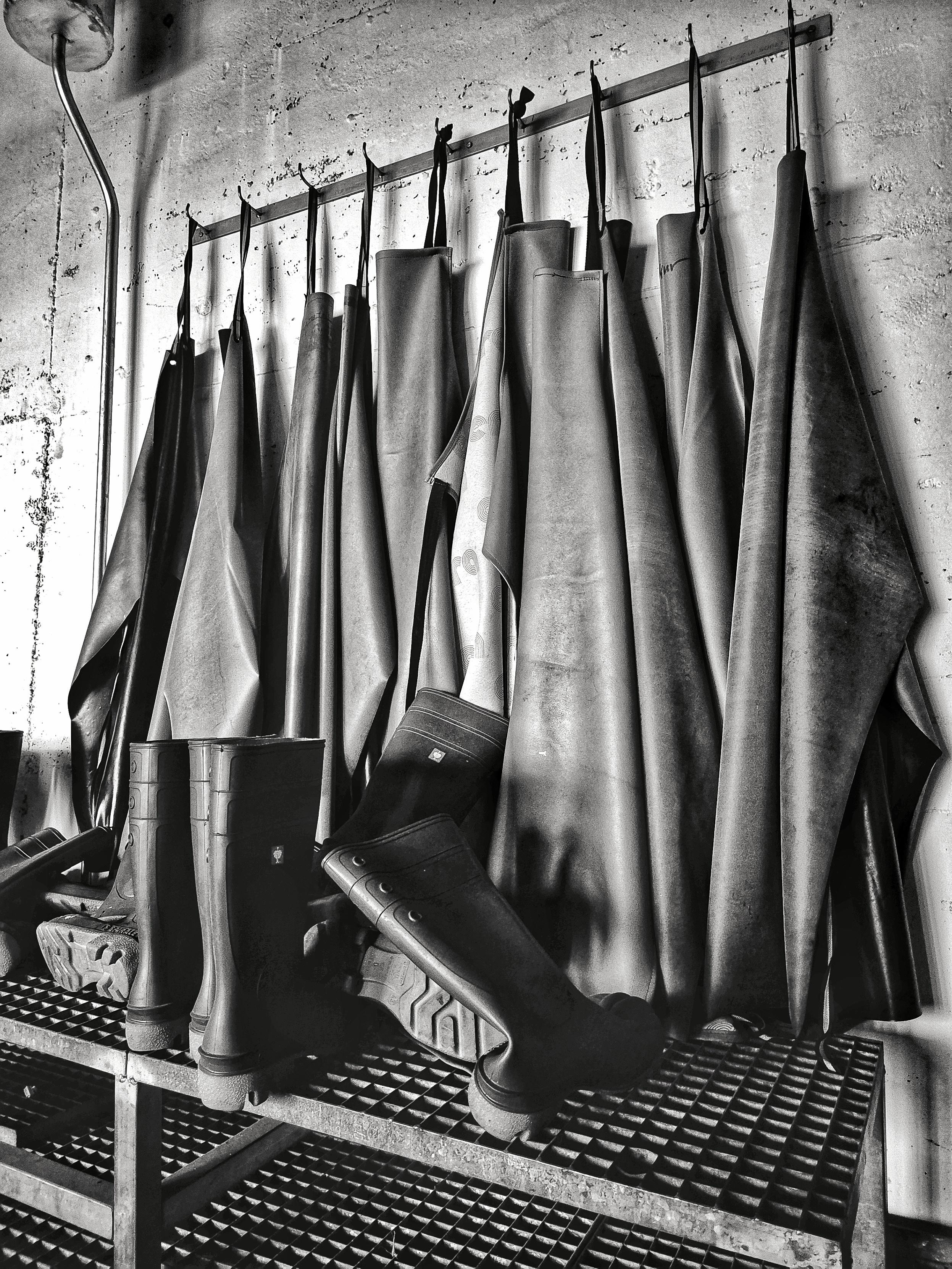 Dieses rechteckige Hochkantfoto in Schwarz-Weiß zeigt eine Reihe von neun Gummischürzen, die an neun Haken einer Metallstange vor einer Betonwand hängen. Darunter stehen auf einem Metallgitterboden mehrere Gummistiefel. Der industrielle und funktionale Eindruck stammt von einem Arbeitsplatz in einer Produktionsstätte.