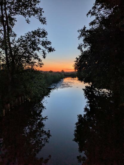 bunter Sonnenuntergang spiegelt sich in einem kleinen Fluß. An der Seite stehen Bäume
