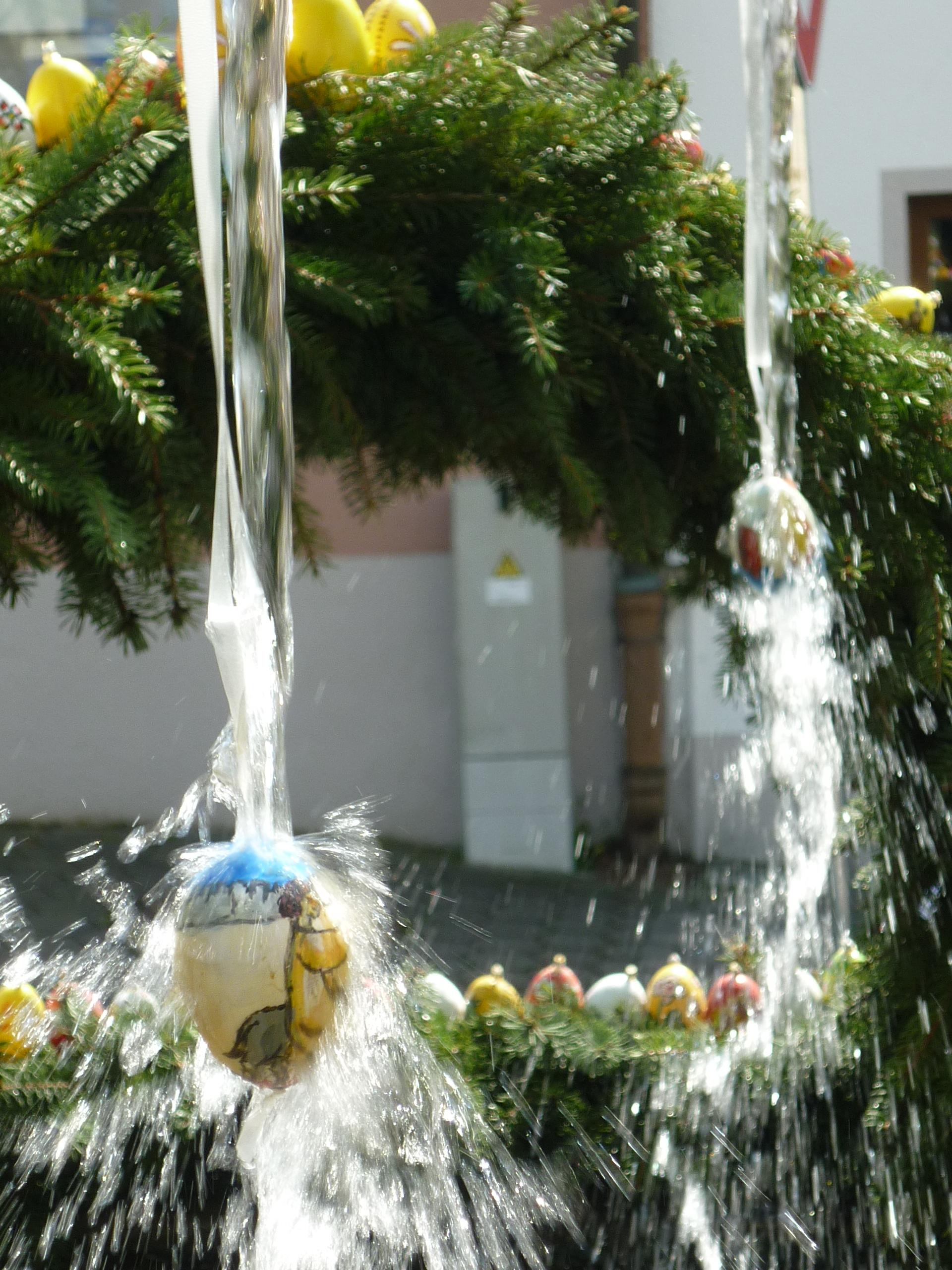 Ausschnitt eines Osterbrunnens. Zwei Ostereier hängen in einem grünen Kranz, über sie läuft Wasser.
