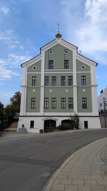 Stirnseite eines Gebäudes, mit zwei weissen Stegen in drei Teile unterteilt.Diese laufen nach oben in den erhöhten Hauptgiebel. Die Unterteilungen sind dunkelgrün gestrichen, die Fenster haben dunkle Rahmen und sind in der Höhe in  Drittel aufgeteilt.