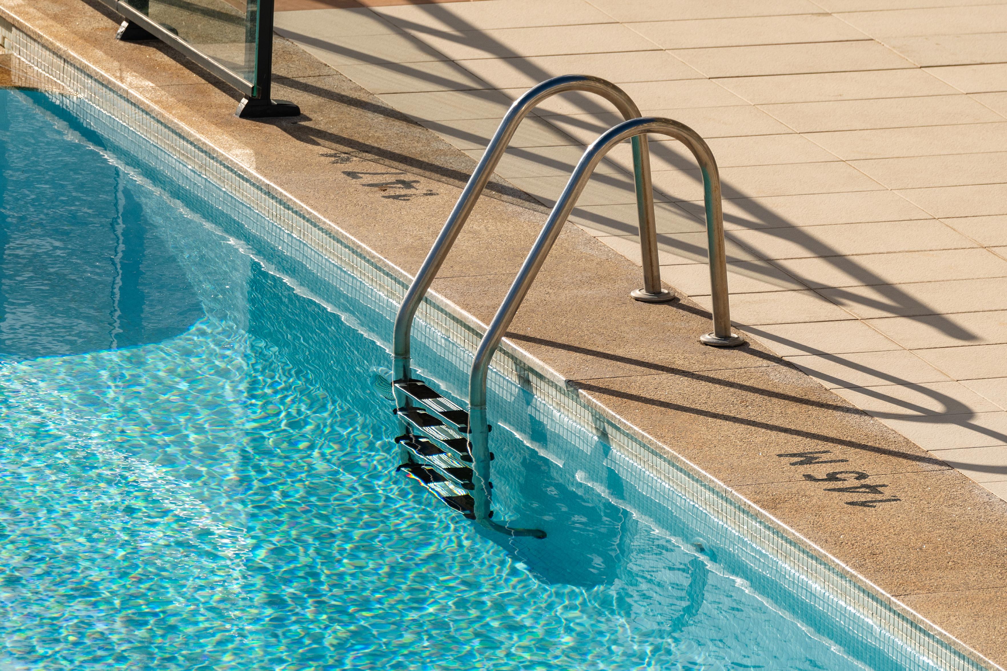 Das Foto zeigt einen Ausschnitt eines Schwimmbeckens mit einer Leiter. Das Wasser ist klar und blau, und die Sonnenstrahlen brechen sich auf der Oberfläche und erzeugen ein glitzerndes Muster. Die Leiter aus Edelstahl ragt aus dem Wasser und führt zum Beckenrand, der mit beigefarbenen Fliesen ausgelegt ist. Auf dem Beckenrand sind Schatten von Geländern zu sehen, die auf eine sonnige Umgebung schließen lassen. Die Markierung "1.45 M" am Beckenrand deutet auf die Tiefe des Wassers hin.