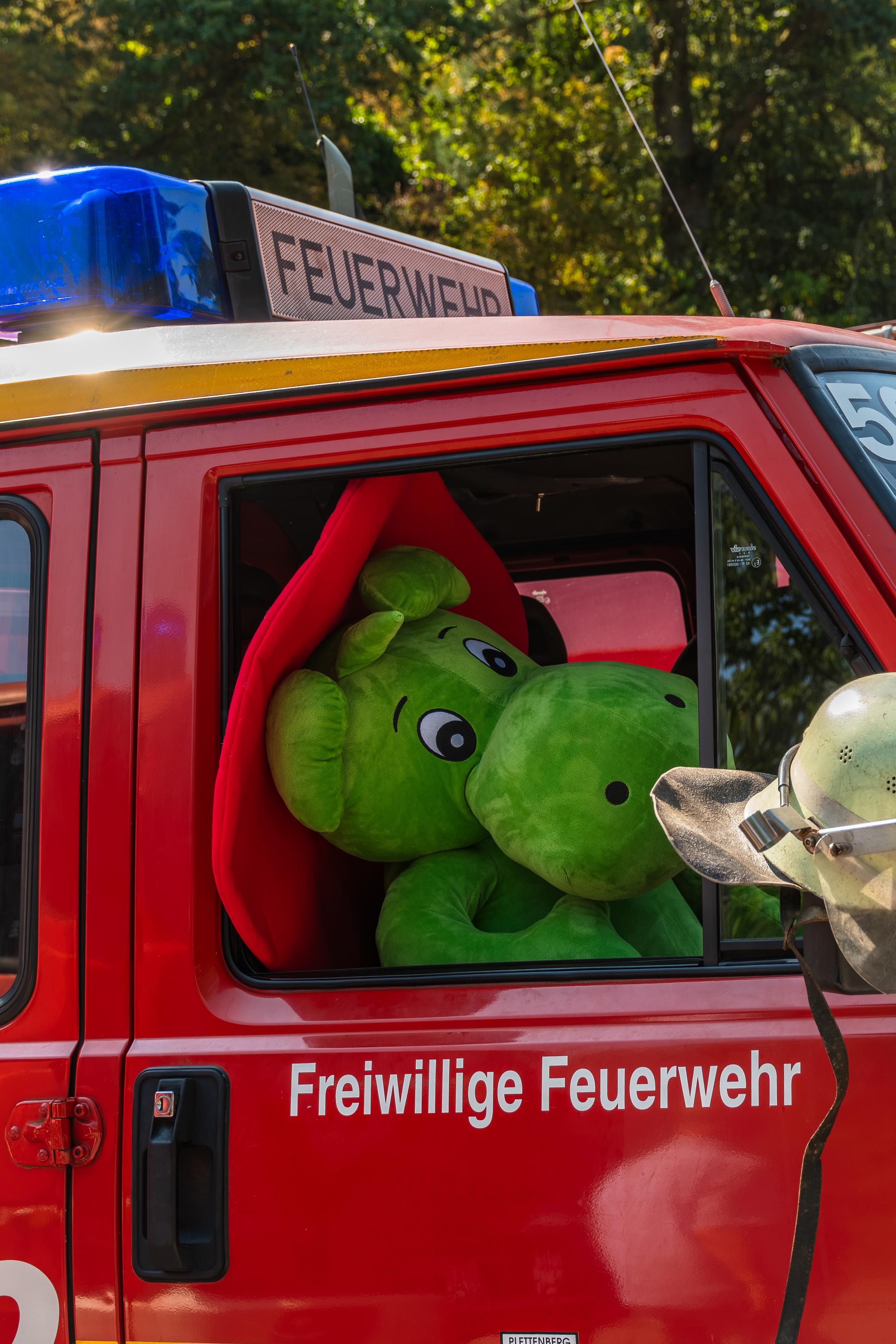 Auf diesem Foto ist ein rotes Feuerwehrauto zu sehen. Auf dem Dach des Fahrzeugs sind blaue Blaulichter und ein Display mit der Aufschrift "FEUERWEHR" angebracht. Durch das Seitenfenster blickt eine grüne, plüschige Figur, die wie ein kleiner Drachen aussieht (bekannt als Grisu), heraus . An der Seite des Feuerwehrautos steht in weißen Buchstaben "Freiwillige Feuerwehr". Daneben hängt ein Feuerwehrmannhelm. Das Bild vermittelt eine fröhliche und verspielte Atmosphäre, die auf eine Veranstaltung oder eine besondere Aktion der Freiwilligen Feuerwehr hindeutet, bei der auch Kinder angesprochen werden sollen.