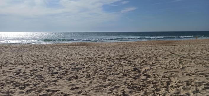 Breiter Sandstrand in ocker mit vielen Fußspuren im Sand, ca 20 Meter zur Wasserlinie und kleinen Wellen. Ins Gegenlicht fotografiert glitzert das Meer auf der linken Seite und spiegelt die Sonne. Rechts steht eine Person am Meer. Himmel ist blau aber leichte Wolken.