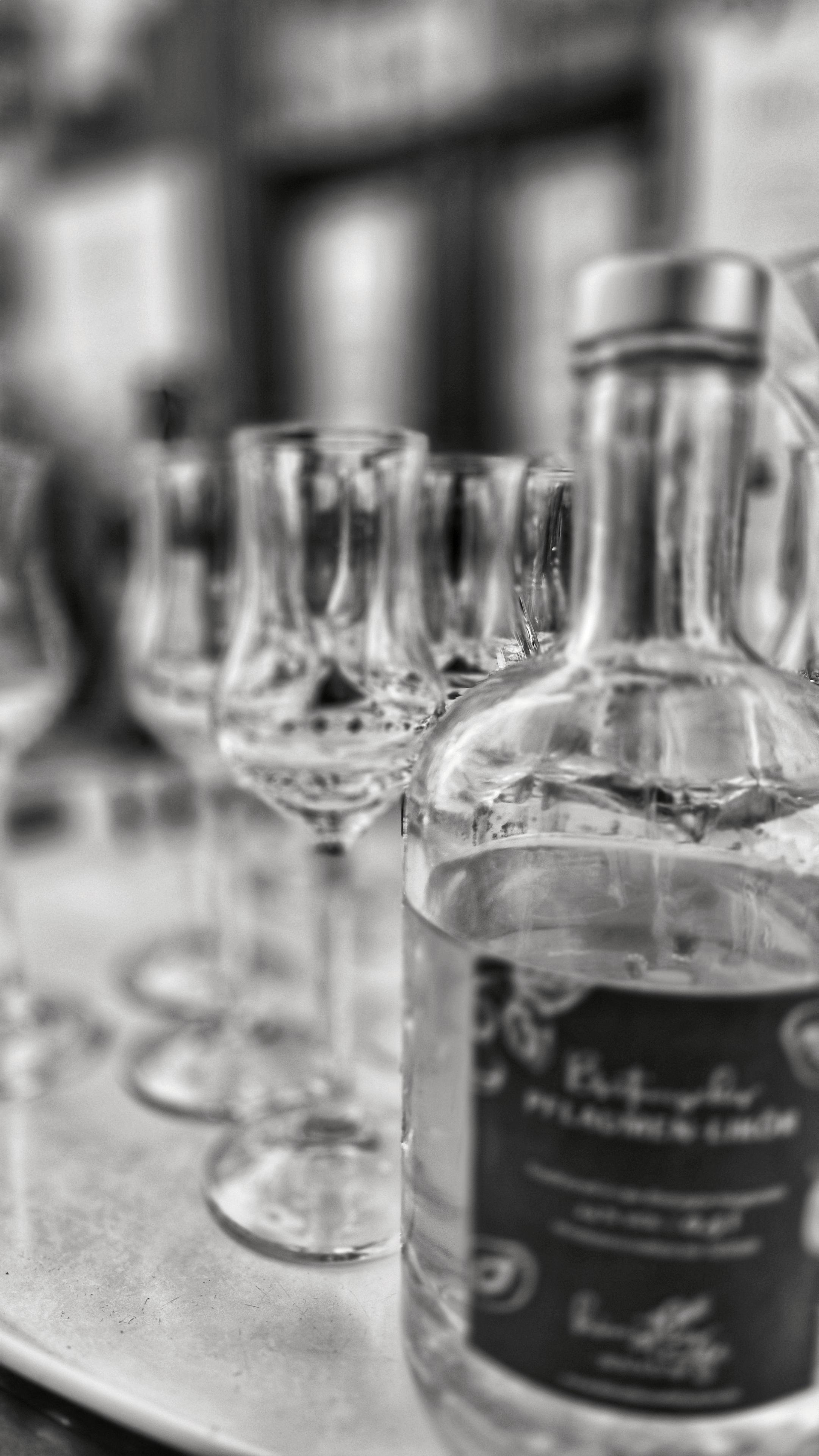 Die Schwarzweiß-Fotografie zeigt eine Flasche mit einem dunklen Etikett im rechten Vordergrund, daneben mehrere kleine Schnapsgläser auf einer Metallplatte. Die Tiefenschärfe ist gering, wobei das vorderste Glas und die Flasche etwas schärfer abgebildet sind, während der Hintergrund verschwimmt. Die Komposition ist vertikal orientiert und verwendet die Gläser als Führungslinie in die Tiefe des Bildes.