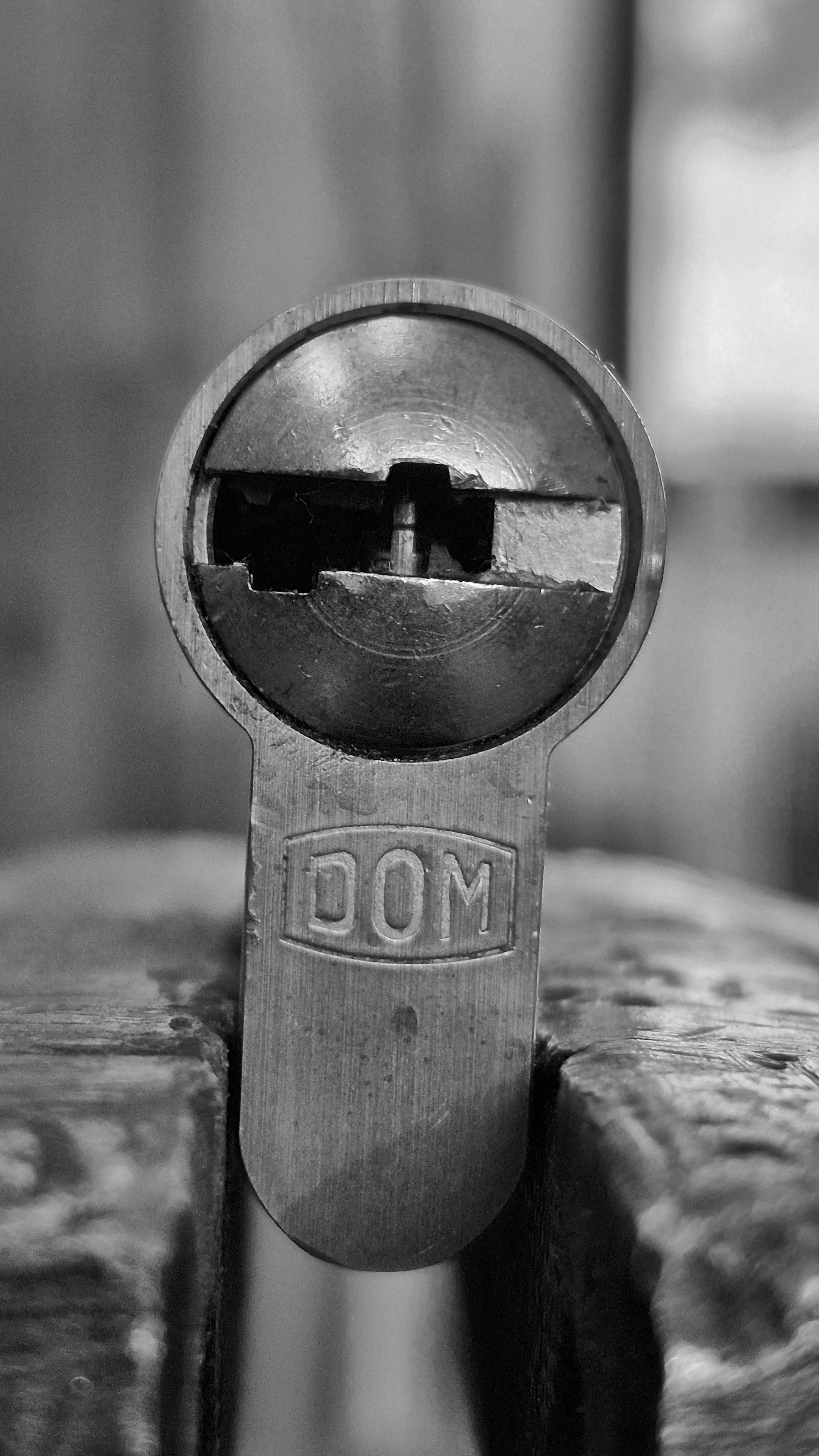 Das Foto ist ein vertikales (hohes) Schwarzweißbild im Hochformat mit einer rechteckigen Form. Es zeigt eine Nahaufnahme eines metallischen Profilzylinders für Türen, der frontal aus unmittelbarer Nähe fotografiert wurde. Der Profilzylinder ist in einen Schraubstock aus Metall eingespannt. Von dem Schraubstock sind allerdings nur die zwei Backen zum Teil zu sehen.
