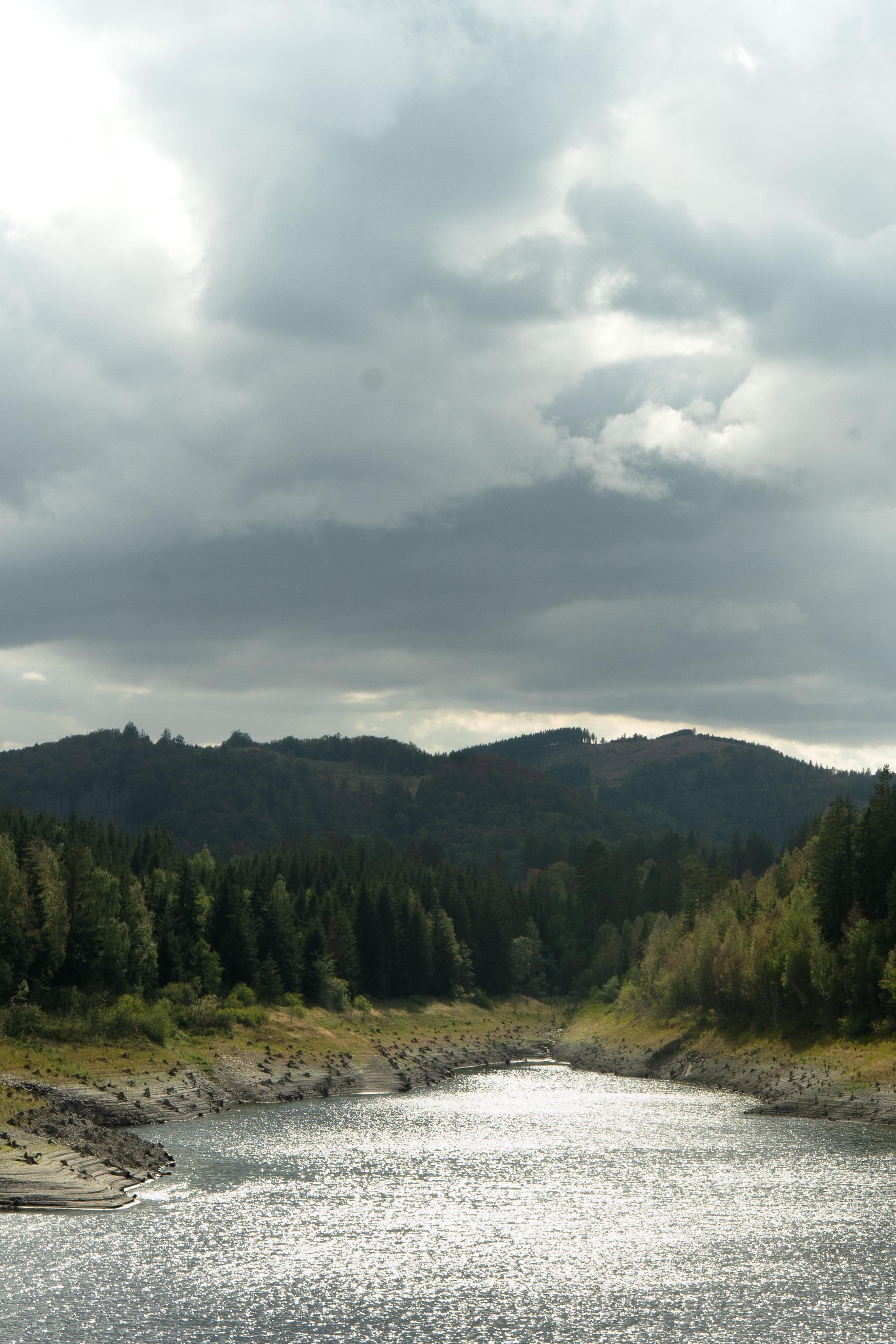 A tranquil river flows through a valley surrounded by dense forests and hills under a cloudy sky. The water reflects a shimmering light, creating a peaceful and serene landscape.