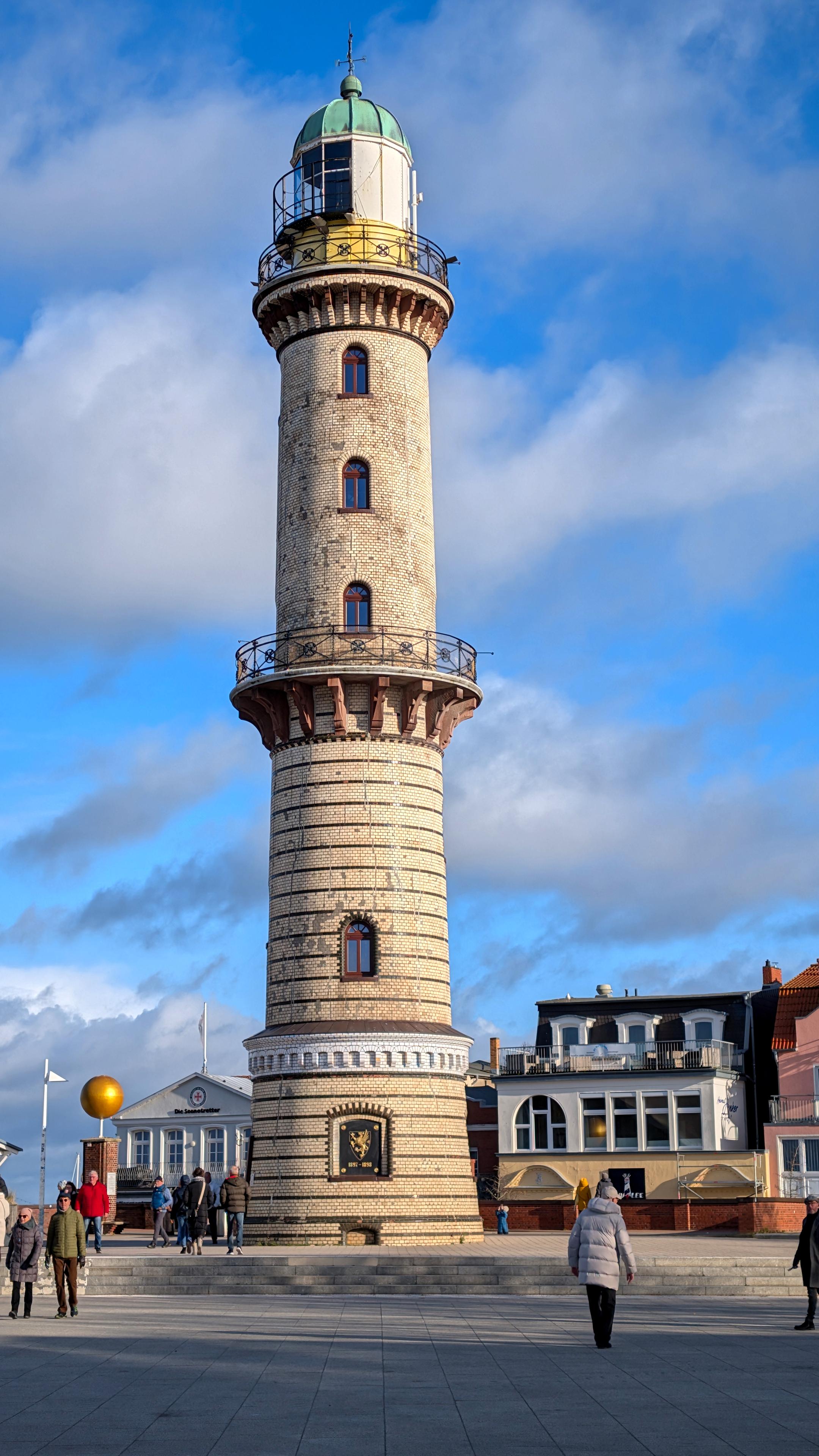 Das Bild zeigt den Leuchtturm von Warnemünde. Der runde Turm zeigt außen helle Ziegel, die unterhalb der ersten umlaufenden Plattform durch einzelne Lagen schwarzer Ziegel unterteilt werden. Oberhalb einer zweiten Plattform steht ein aus Metall errichteter zylindrischer Aufsatz, der die Leuchte enthält. Er ist zur See hin verglast und besitzt oben eine grüne Kuppel mit Wetterkreuz.
Auf dem Platz davor laufen Menschen herum. Im Hintergrund Häuser. Blauer Himmel mit weißen Wölkchen.