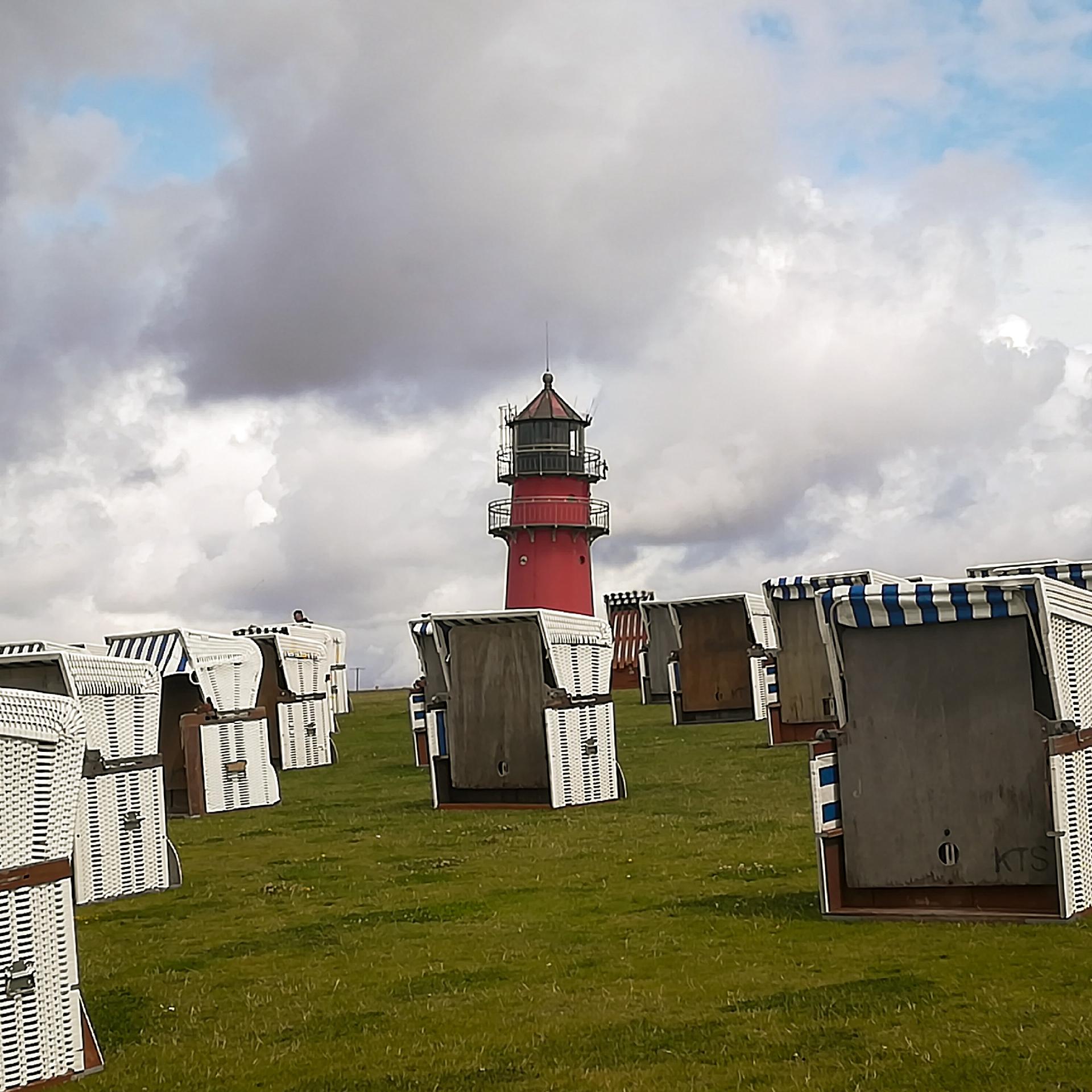 Büsumer Leuchtturm in Rot, halb hinter einem grünen Wiesenhügel versteckt, auf dem weiße Strandkörbe stehen.